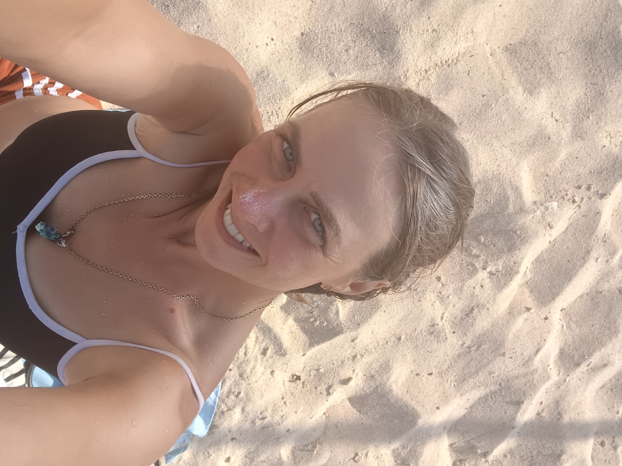 A woman lies on a sandy beach, her body angled to the left as she takes a selfie. Her hair is damp and pulled back, framing a face that smiles at the camera. She wears a black bikini with white trim and a silver chain necklace with a blue pendant. The sand is light brown and textured, with shadows cast across it, suggesting an outdoor setting. The scene is likely located on a beach in Nha Trang, Vietnam.