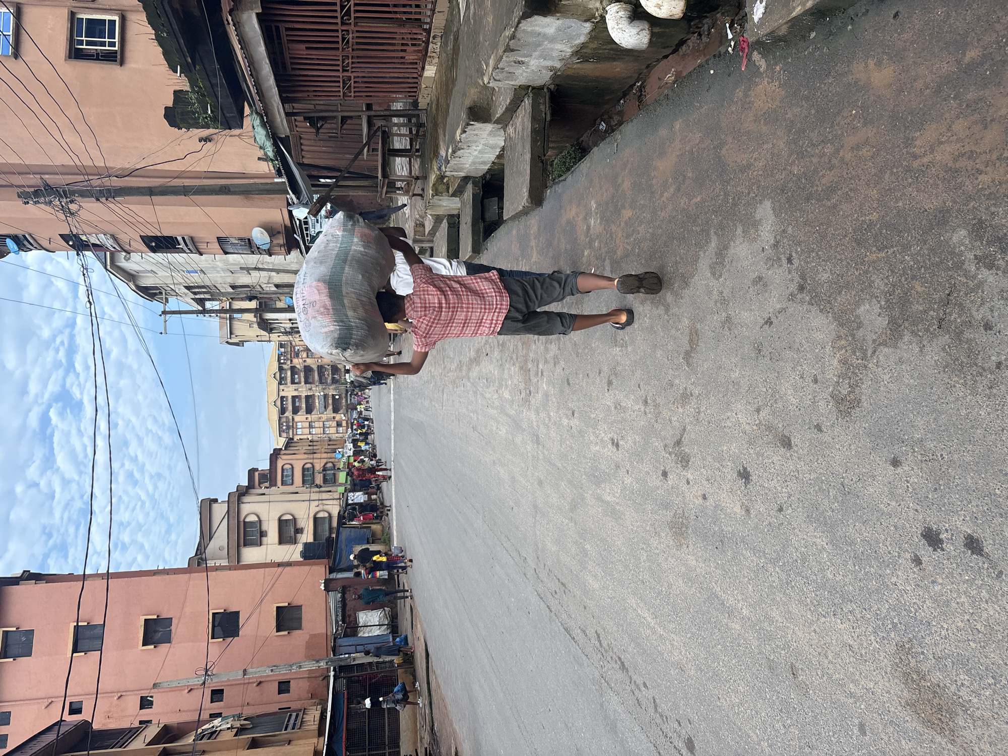 This image depicts a street scene in Onitsha, Nigeria. The foreground is dominated by an asphalt road with a concrete sidewalk on the right. A young boy wearing a red and white checkered shirt and grey shorts walks away from the viewer, carrying a large, bundled sack on his head. He is accompanied by another person, partially visible, walking in the same direction.
The street is lined with multi-story buildings on both sides. The buildings exhibit a range of architectural styles and states of repair, with some appearing weathered and others more modern. Power lines crisscross the sky above the street. On the left side of the street, various structures, some with open fronts resembling market stalls, are visible. People are scattered along the street and sidewalk, some appearing to be engaged in activities related to trade or daily life. The sky is partly cloudy. The overall impression is of a bustling urban environment.
