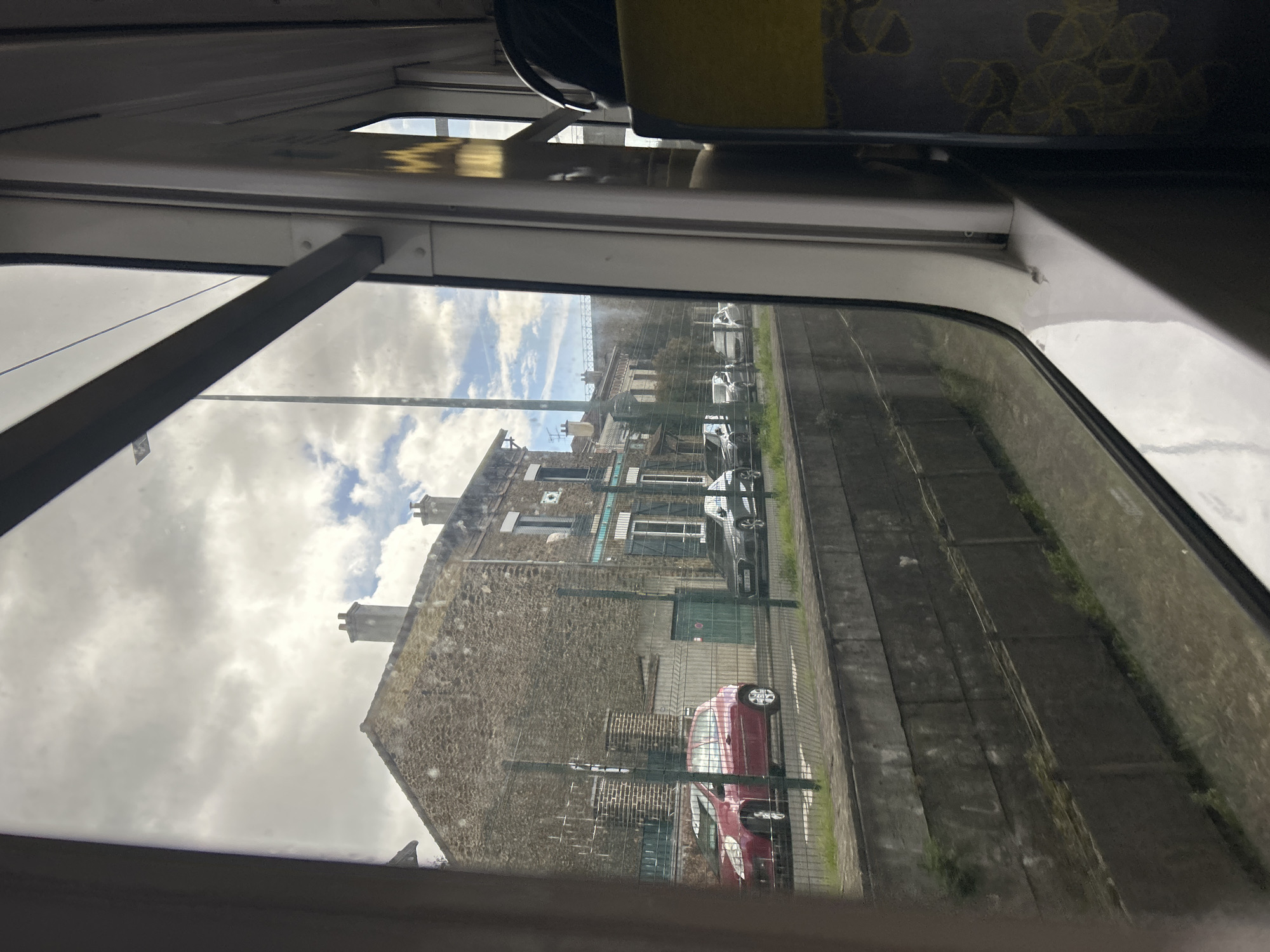 The image depicts an exterior urban scene in Arcueil, France, observed from inside a vehicle, likely a train or tram, through a window. The immediate foreground shows the white interior window frame and a portion of a yellow and dark grey patterned seat on the right. Reflections are visible on the window glass.
Outside the window, a concrete canal or trench with weathered grey walls, partially covered by sparse green vegetation, runs horizontally across the lower third of the frame. Above this, a green metal fence separates the canal area from a paved street. Several vehicles are parked along this street. Prominently, a red three-door hatchback is parked closest to the viewer, followed by a dark grey sedan or hatchback further down the street. Additional vehicles, lighter in color, are visible in the distance.
In the background, a row of multi-story buildings with stone facades, pitched roofs, and multiple chimneys lines the street. The most prominent building on the left features rough-hewn stone and a dark roof, while an adjacent building to its right has blue decorative trim around its windows and dark shutters. The sky overhead is partially overcast with large grey clouds interspersed with patches of blue sky and white cumulus clouds. A tall, slender utility pole extends vertically into the sky on the right side of the main building. No individuals are visible. The scene is static, captured from a moving perspective.