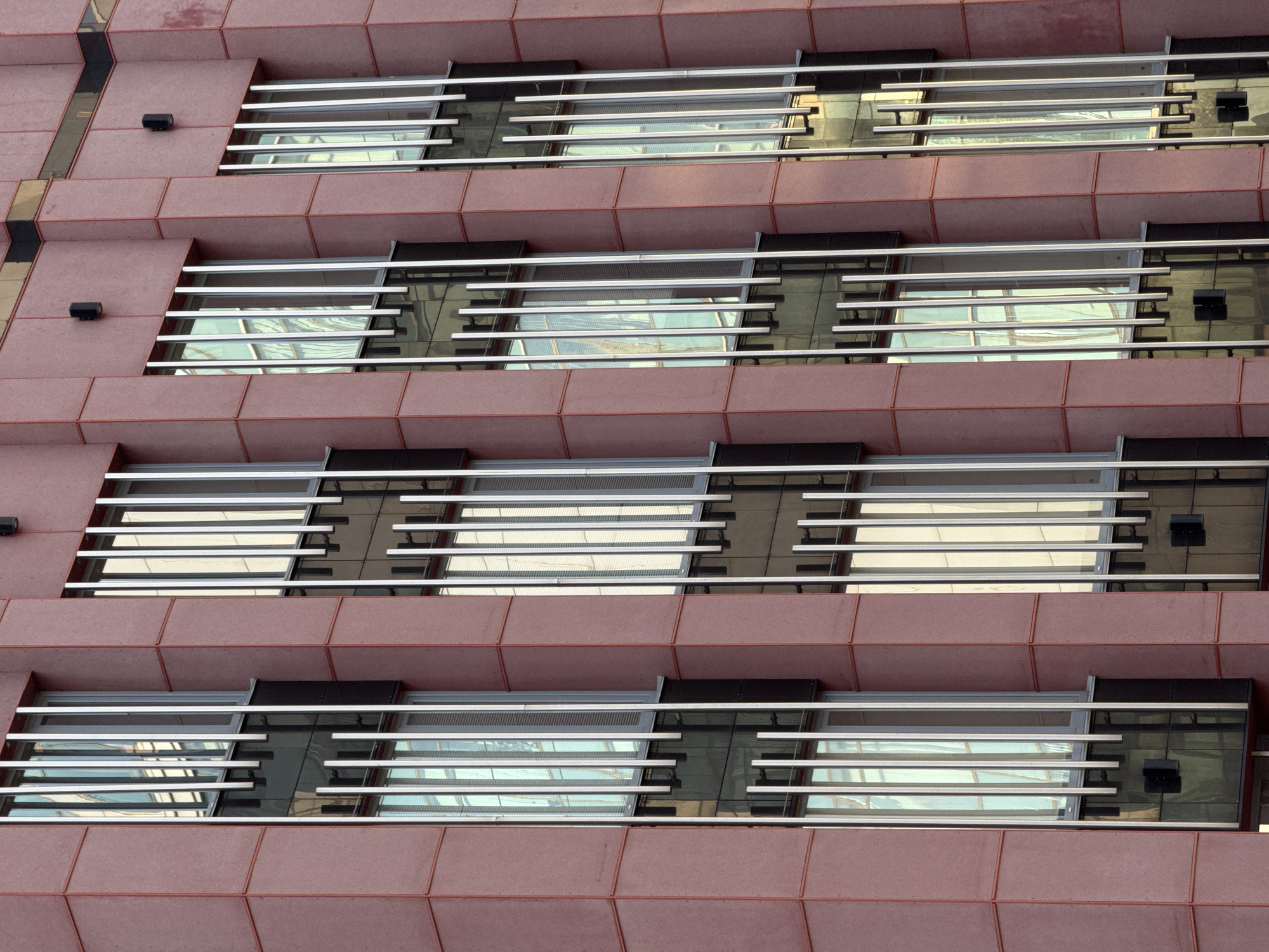 This media file is a close-up, eye-level shot of the exterior of a modern building, likely an office or residential building, in Izhevsk, Russia. The building's facade is constructed with large, rectangular panels of a reddish-pink material, possibly concrete or stone. Interspersed between these panels are horizontal bands of windows, each protected by a series of thin, horizontal silver-colored louvers.
The windows themselves are dark and reflective, showing the sky and the surrounding environment, though the specific details of the reflection are somewhat abstract due to the angle and the louvers. The louvers are positioned at an angle, creating a layered effect and partially obscuring the view into the building. Some of the windows appear to have small black boxes attached to their exterior, which could be security cameras or lighting fixtures.
The repeating pattern of the panels and the windows creates a sense of rhythm and order. The lighting suggests it is daytime, but the sky appears overcast, as there are no strong shadows. The overall impression is one of architectural design focused on clean lines and geometric shapes, with a utilitarian yet aesthetically considered approach to fenestration and facade treatment. There is no visible text in the image.