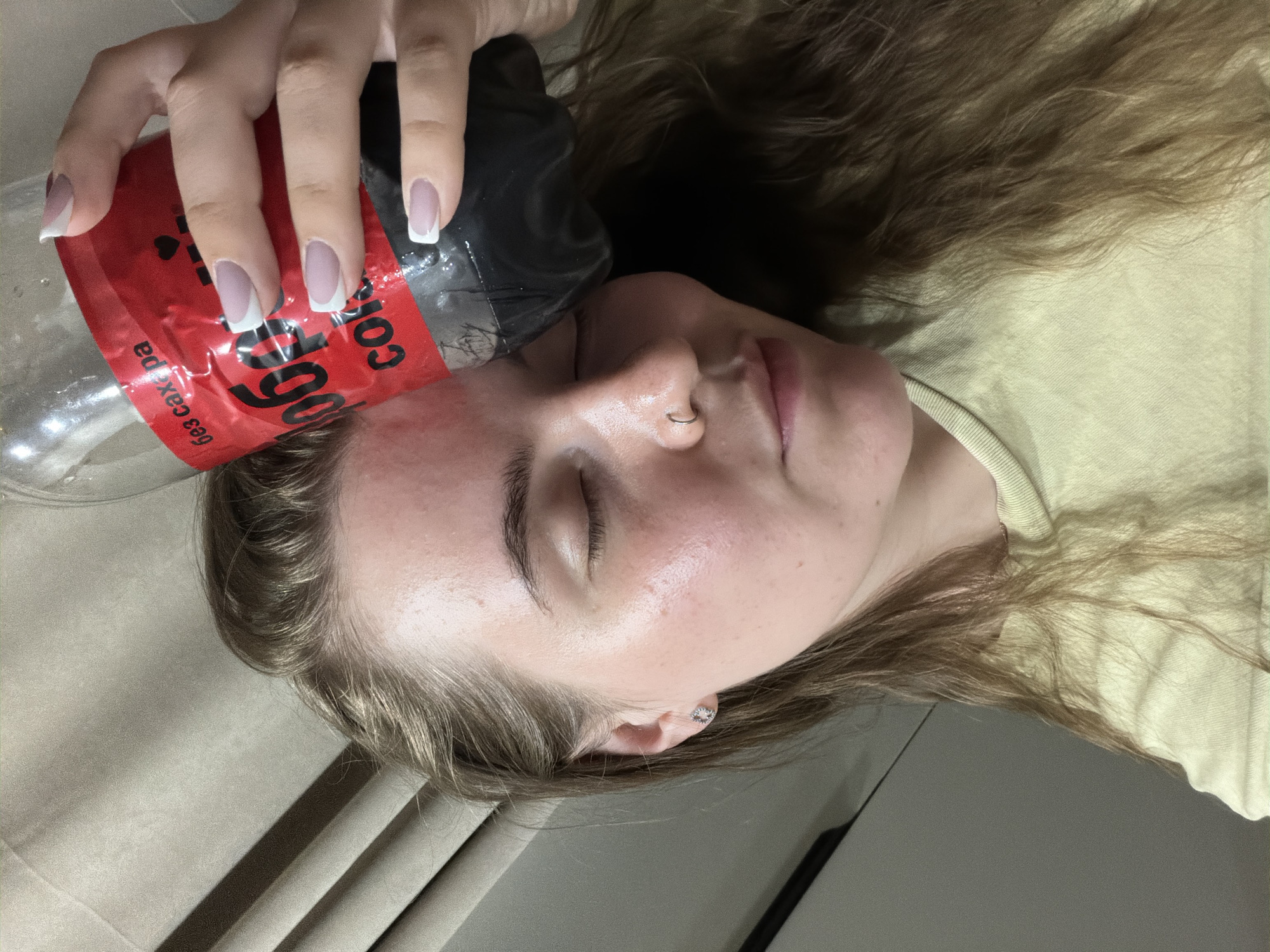 A woman is lying on her back with her eyes closed, holding a red and black "Coca-Cola" branded bottle against her left cheek. She has a nose ring in her left nostril and an earring in her left ear. Her hair is spread around her head. The bottle is partially empty. The setting appears to be indoors, with draped fabric visible beneath her and a light-colored garment visible on her torso. The language printed on the bottle is Cyrillic, indicating that the photo may be from Russia. The "Coca-Cola" label includes the wording "без сахара" (bez sakhara) which translates to "sugar-free" or "zero sugar." The bottle also has "1006" written on it. There is no contextual information within the image to verify it was taken in Chaikovsky, Russia.
