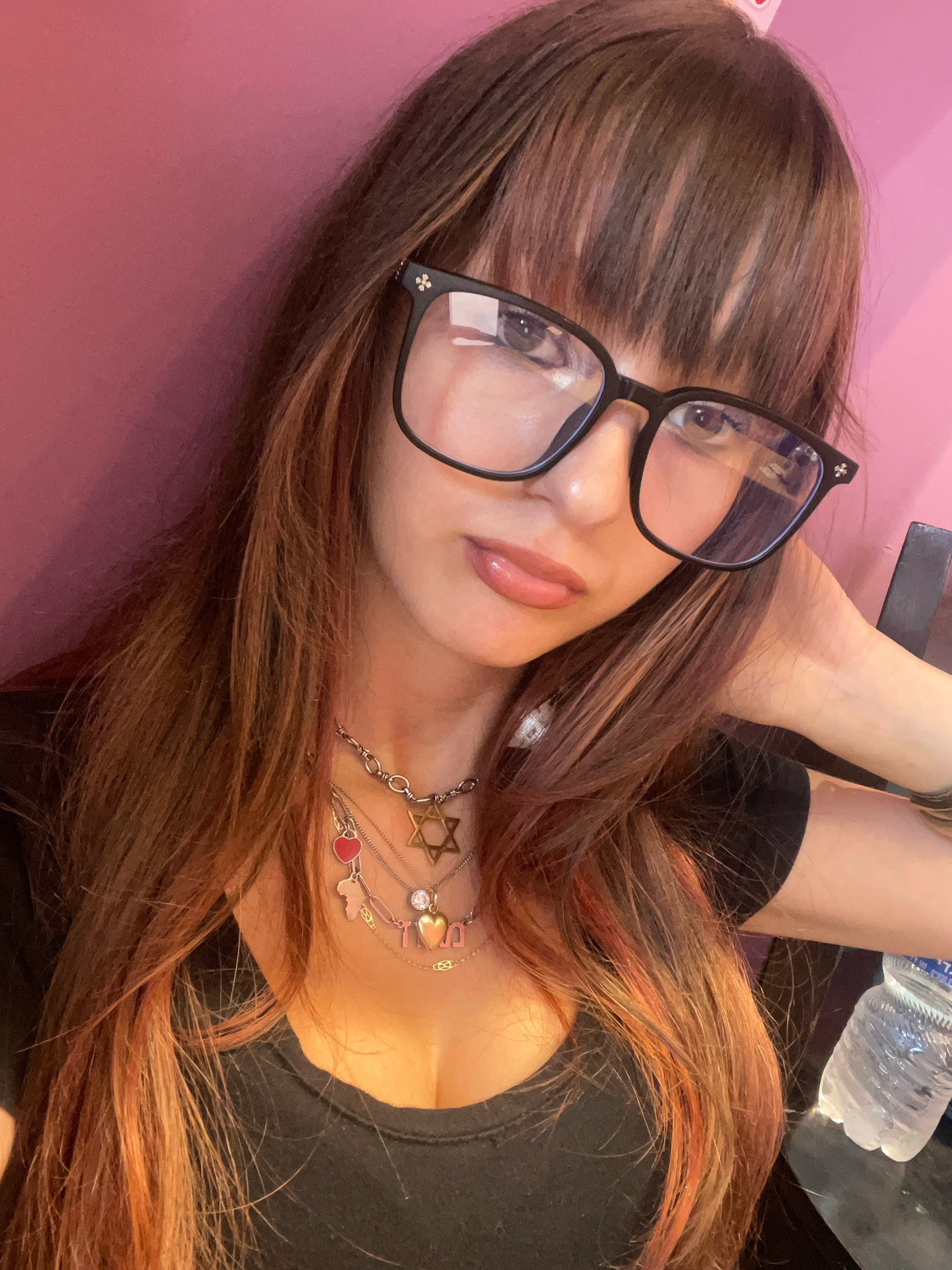 A medium shot photograph taken indoors shows a young woman with long, dark hair with red highlights, wearing black-rimmed glasses and a black t-shirt. She is wearing several necklaces, including one with a Star of David pendant and a heart pendant. Her left arm is resting behind her head, and her right hand is on her lap, near a bottle of water. The background is a pink wall. There is no specific indication of the city of Raanana, Israel, in the image.