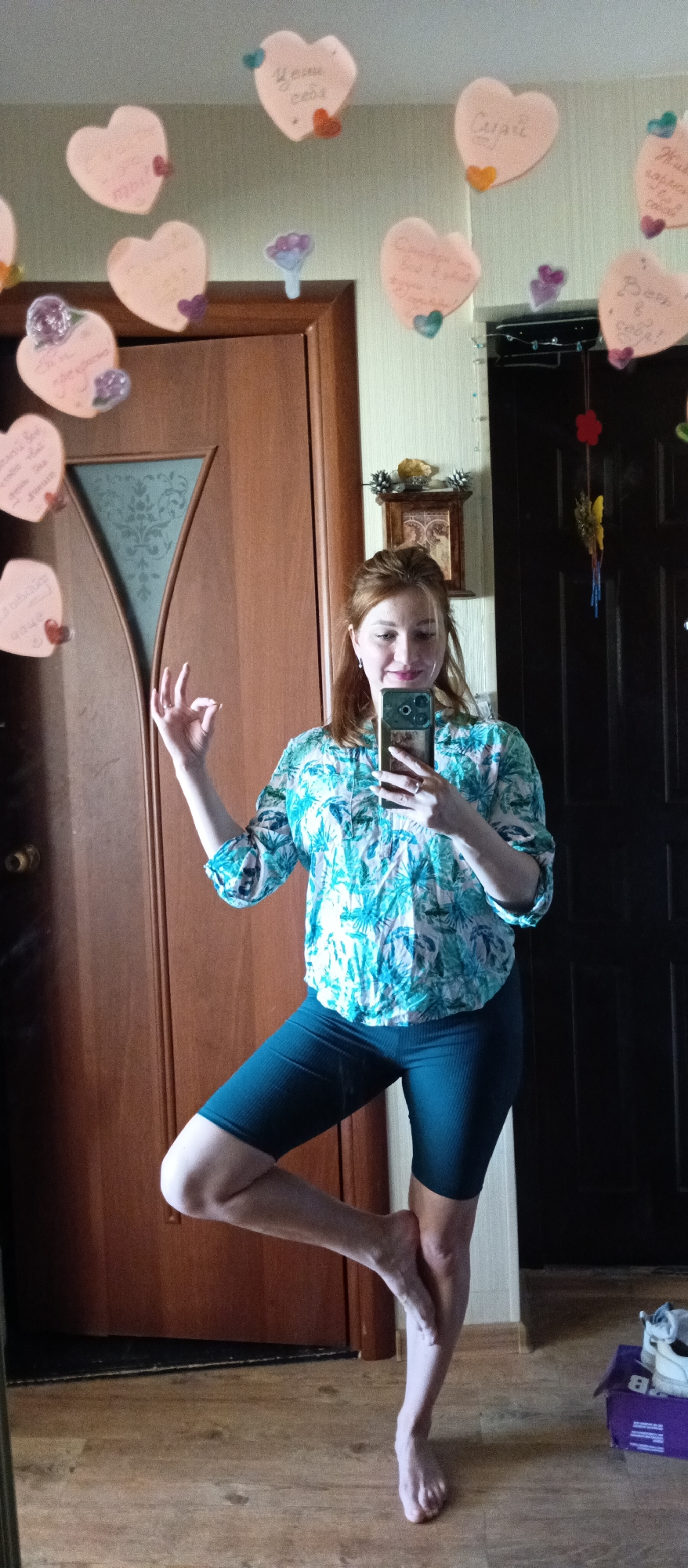 A woman with reddish-brown hair is standing on one leg, with the other foot placed against her inner thigh, in front of a mirror. She is wearing a blue patterned blouse and dark green shorts. She is holding a smartphone and taking a selfie. Her left hand is raised, forming an "okay" sign. The mirror has paper hearts with text on them attached to it. Behind her are two doors, one brown and one dark gray. The floor is wooden. There are some objects on the floor near the bottom right of the image. The location is indoors, likely in a home.
