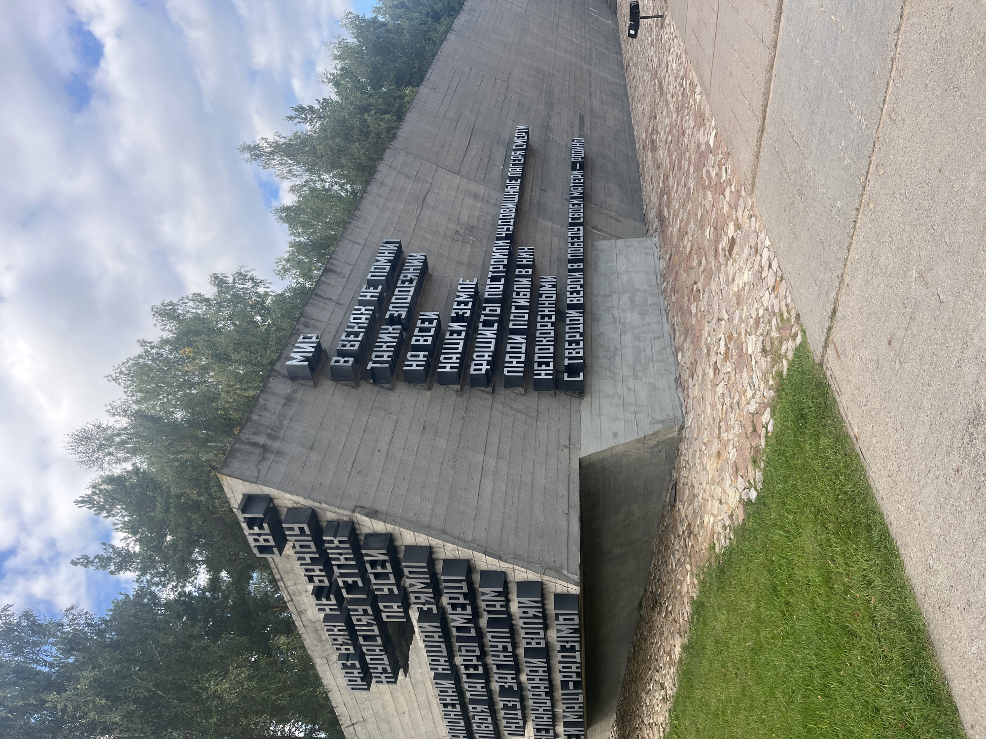 The image depicts a concrete monument with raised black letters in Cyrillic script, located in a park-like setting. The monument is a long, angular structure, with the text arranged in several lines. The left side of the monument features text in Belarusian, with phrases such as "ПРАЎВАГІ НЕ БАЧЫЦЬ", "НАСУСЯЧ ГЭТКІХ", "ПА УСЕЙ", "НАШЫЯ ЗЯМЛІ", "ЛЮДЗІ ЗАГНУЛІ ТАМ", "НЕПАКОРАНЫМ ВОПЛЯМ", and "СЯ МАЦІ-РАДЗІМЫ". The right side of the monument displays text in Russian, including "МИР", "В ВЕКАХ НЕ ПОМНИТ", "ТАКИХ ЗЛОДЕЯНИЙ", "НА ВСЕЙ", "НАШЕЙ ЗЕМЛЕ", "ФАШИСТЫ ПОСТРОИЛИ ЧУДОВИЩНЫЕ ЛАГЕРЯ СМЕРТИ", "ЛЮДИ ПОГИБЛИ В НИХ", "НЕПОКОРЕННЫМИ", and "С ТВЕРДОЙ ВЕРОЙ В ПОБЕДУ СВОЕЙ МАТЕРИ – РОДИНЫ".
The monument is situated on a paved area, bordered by a strip of lush green grass on the left and a path made of irregularly shaped stones behind it. In the background, tall, green trees line the horizon under a sky with scattered white clouds. A small black object, possibly a trash receptacle or a plaque, is visible to the right of the monument near the stone path. The overall scene suggests a memorial site, likely commemorating victims of war or persecution. The monument's design is stark and imposing, with the bold lettering emphasizing the solemnity of the message.