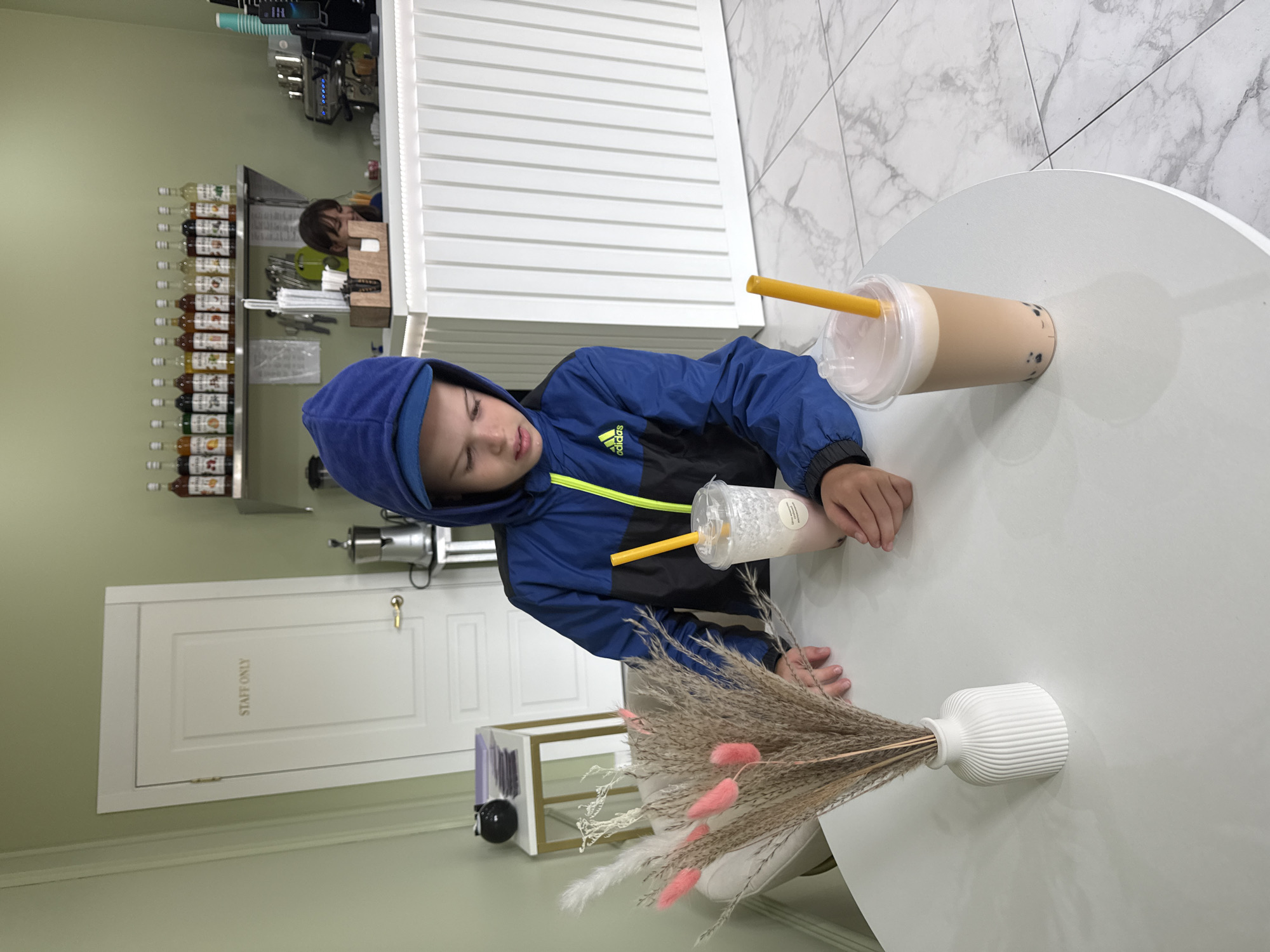 A young boy wearing a blue Adidas hooded jacket with a yellow zipper is sitting at a white round table in a modern cafe. He is looking down at a plastic cup with a pinkish beverage and a yellow straw. In the foreground on the table, a white ribbed vase holds a bouquet of dried flowers, including some pink bunny tail grass. Another identical plastic cup with a light brown beverage and a yellow straw is to the right of the boy. In the background, a white counter with a fluted design spans across the space. Behind the counter, a woman with dark hair is visible, smiling and seemingly working. A shelf above the counter holds numerous bottles of flavored syrups. To the left of the boy, a white door is visible with "STAFF ONLY" written in gold letters. The cafe has light green walls and a marble-patterned tiled floor. The overall impression is a bright and contemporary cafe setting. The location is not specified in the image.