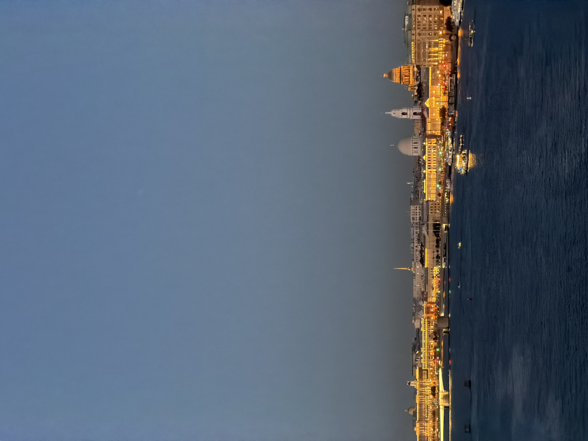 This is a wide-angle, vertical, outdoor, eye-level, full-shot photograph taken at dusk over the Neva River in Saint Petersburg, Russia. The sky is a deep, clear blue, transitioning to a darker gradient near the horizon. The cityscape of Saint Petersburg stretches across the middle ground, illuminated by warm, golden lights. Prominent structures include several domed buildings, including what appears to be St. Isaac's Cathedral on the right, identifiable by its large golden dome. Other buildings feature neoclassical architecture with numerous windows. A long bridge spans the river, with traffic lights visible on its deck. Several boats and ships are navigating the dark blue water of the Neva River, their lights reflecting on the surface. The overall scene depicts a serene twilight view of the historic city.