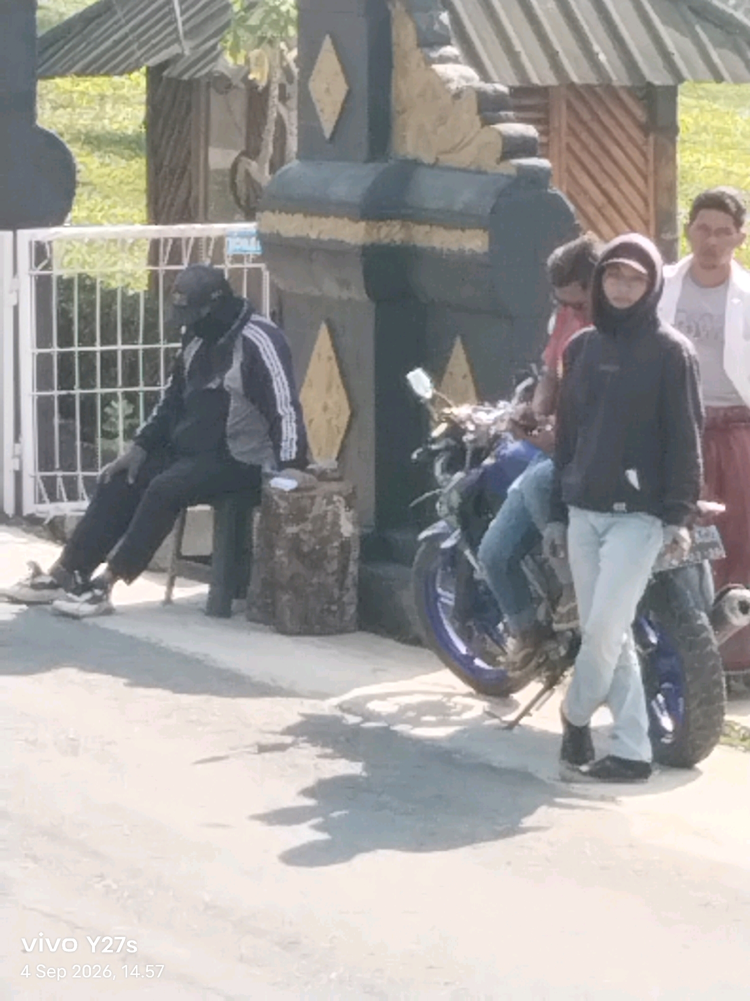In Sigedang, Indonesia, a man wearing a dark hoodie and light blue jeans stands on the right side of the frame near a motorcycle. To his left, another person wearing a dark jacket with white stripes and dark pants sits on a stool. This individual has a dark cap covering their head and a dark mask obscuring their face. A blue motorcycle is parked in the center of the image. In the background, a partially visible black and gold structure stands, with a white metal fence to the left and greenery beyond. A white shirt and a light-colored garment are visible on two other individuals further in the background. The ground is paved, and shadows are cast across it. The timestamp "vivo Y27s 4 Sep 2026, 14.57" is visible in the lower left corner.