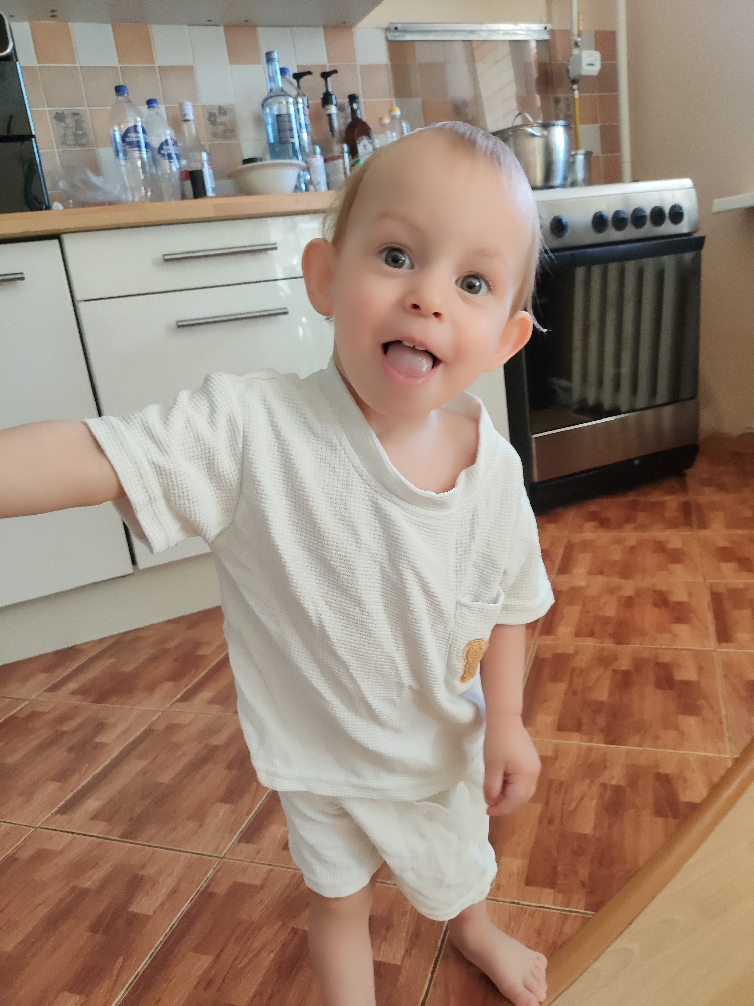 A young child in a white t-shirt and white shorts stands on a brown-tiled floor in a kitchen setting. The child has fair skin, light-colored hair, and is sticking their tongue out with a playful expression. Kitchen cabinets and appliances, including an oven and bottles on the counter, are visible in the background. The location is Chaikovsky, Russia.