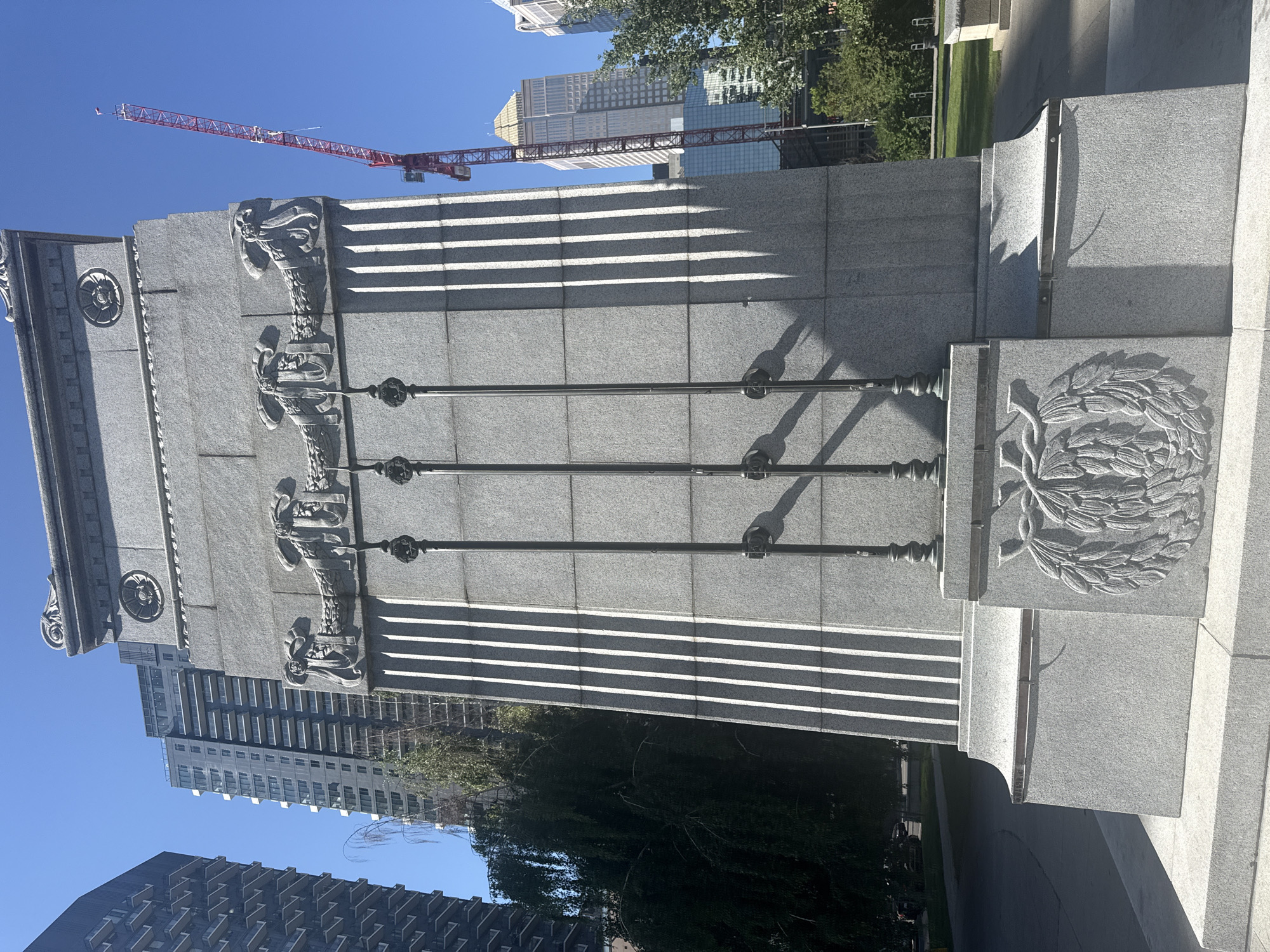 The image shows a large granite monument in a city park, with modern high-rise buildings and a construction crane visible in the background. The monument is rectangular and features fluted columns, decorative carvings of laurel wreaths and floral motifs, and three tall, slender poles extending upwards. Sunlight casts distinct shadows across the monument's surface. The environment is urban, with a clear blue sky overhead. This monument is located in Olympic Plaza in Calgary, Alberta, Canada.