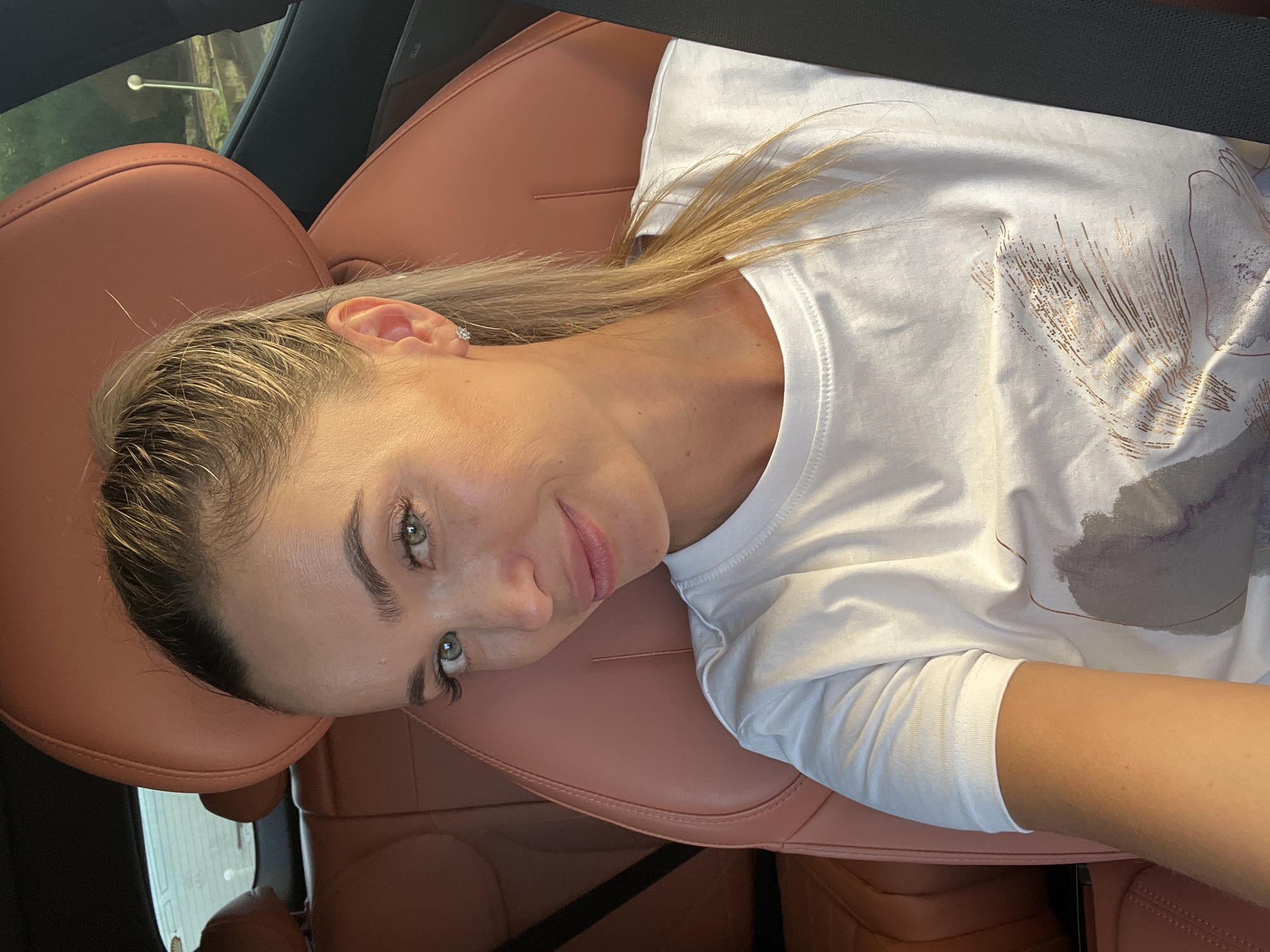 A young woman with blonde hair pulled into a ponytail and green eyes is sitting in the driver's seat of a car. She is wearing a white t-shirt with a graphic design on it. The car has tan leather seats. The background shows the interior of the car and some greenery and a street light outside the window.