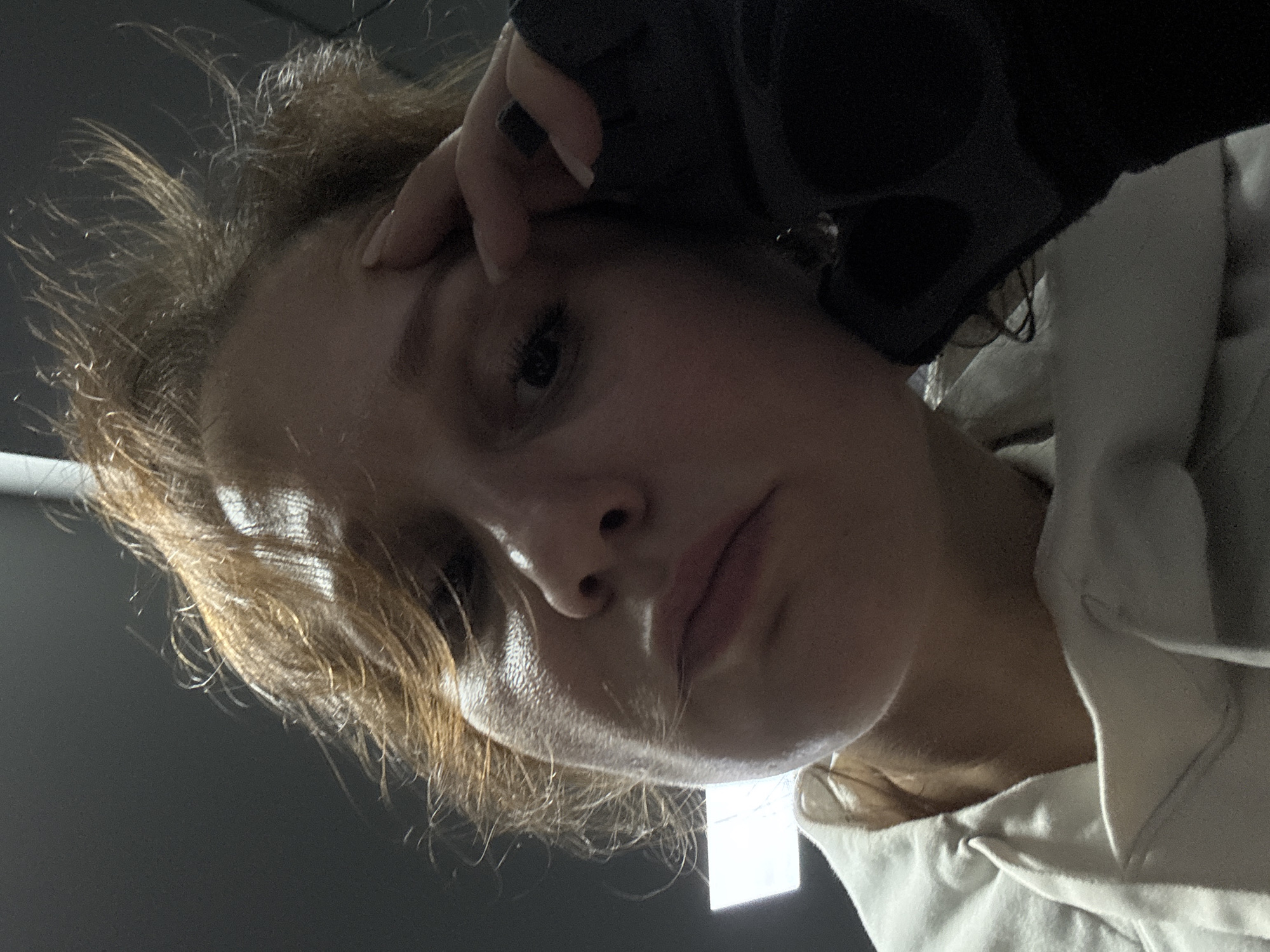 A close-up, angled downward shot shows a light-skinned person with short, tousled, light brown hair. The person's right hand, clad in a black glove or covering, is touching their forehead. The person has dark eyes and slightly parted lips. They are wearing a white hooded garment. The background appears to be a dark grey or black. There is a bright light source partially visible at the bottom of the frame. There is no information in the image to suggest the location is Dubai, United Arab Emirates.
