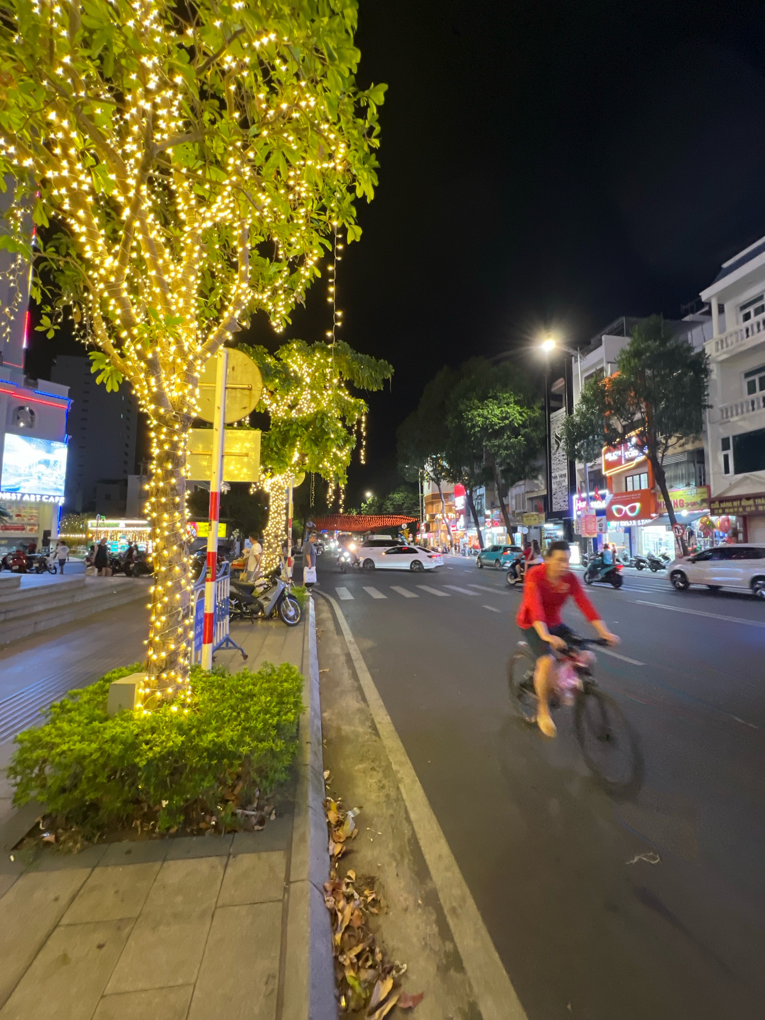 This is a nighttime photograph taken on a street in Nha Trang, Vietnam, likely a commercial or pedestrian area. The scene is illuminated by numerous string lights decorating trees along the sidewalk, casting a warm glow. In the foreground, a tree wrapped in yellow fairy lights dominates the left side of the frame, with a manicured green hedge at its base. A sidewalk, paved with large rectangular tiles, runs alongside the street.
On the street, a person is riding a bicycle, blurred due to motion. They are wearing a red shirt and dark shorts, with a pink bag attached to the handlebars. Several other vehicles, including motorcycles and cars, are visible on the road, some of which are also blurred. Buildings line the right side of the street, displaying illuminated signs and storefronts. The architecture appears to be a mix of modern and older styles. The overall atmosphere suggests a vibrant urban environment at night, possibly a popular spot for locals and tourists.