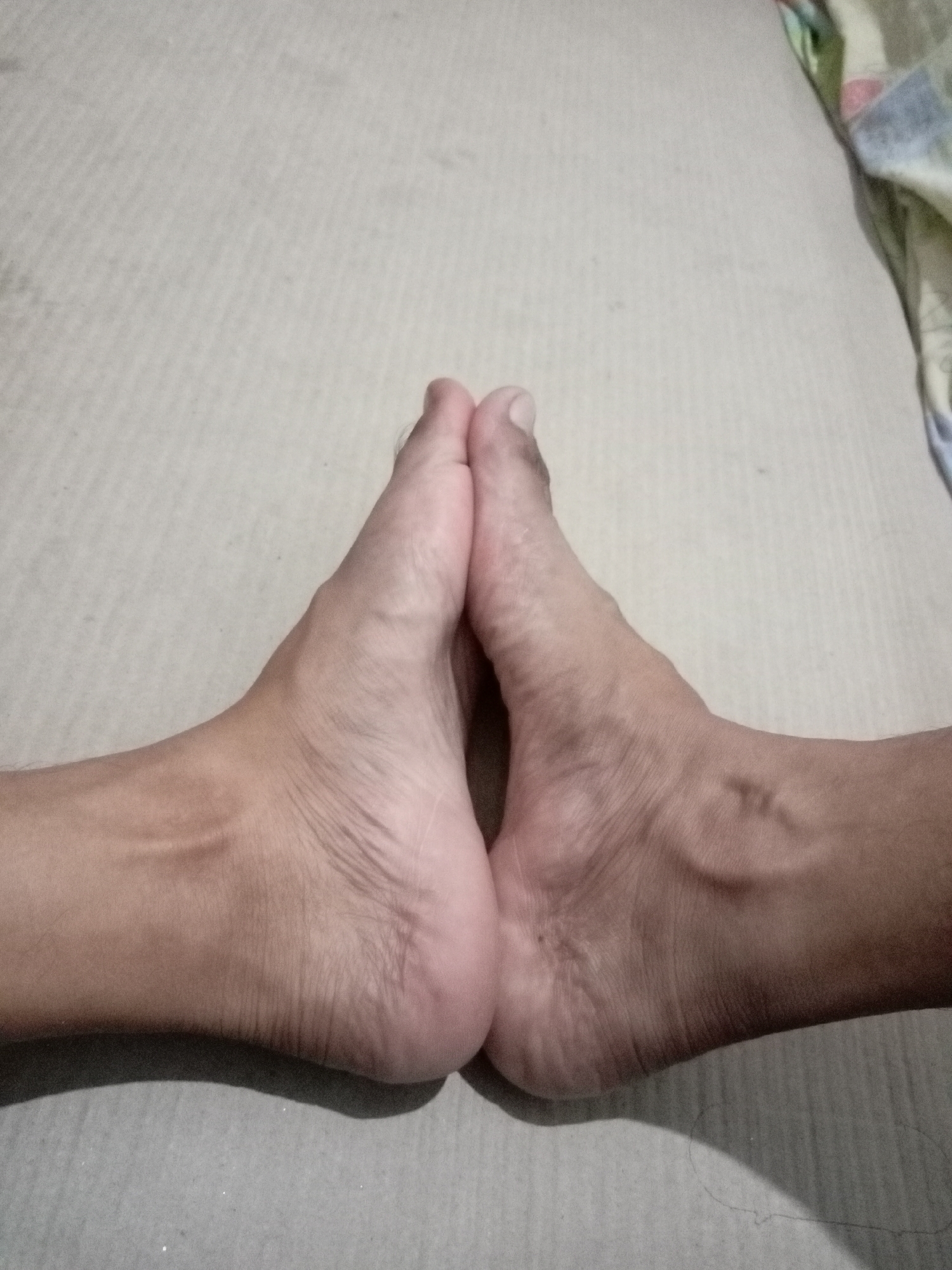 This image shows a close-up view of two bare feet positioned together, soles facing each other, on a textured, light-colored surface. The feet appear to be resting on a bed or mattress, as indicated by the fabric visible on the right side of the frame, which has a subtle patterned weave and appears to be a corner of bedding with multi-colored stripes. The feet exhibit visible veins and skin creases, characteristic of adult feet. The lighting casts a soft shadow beneath the feet. The image quality is slightly grainy. No specific location in Batam, Indonesia, can be determined from the visual information presented.