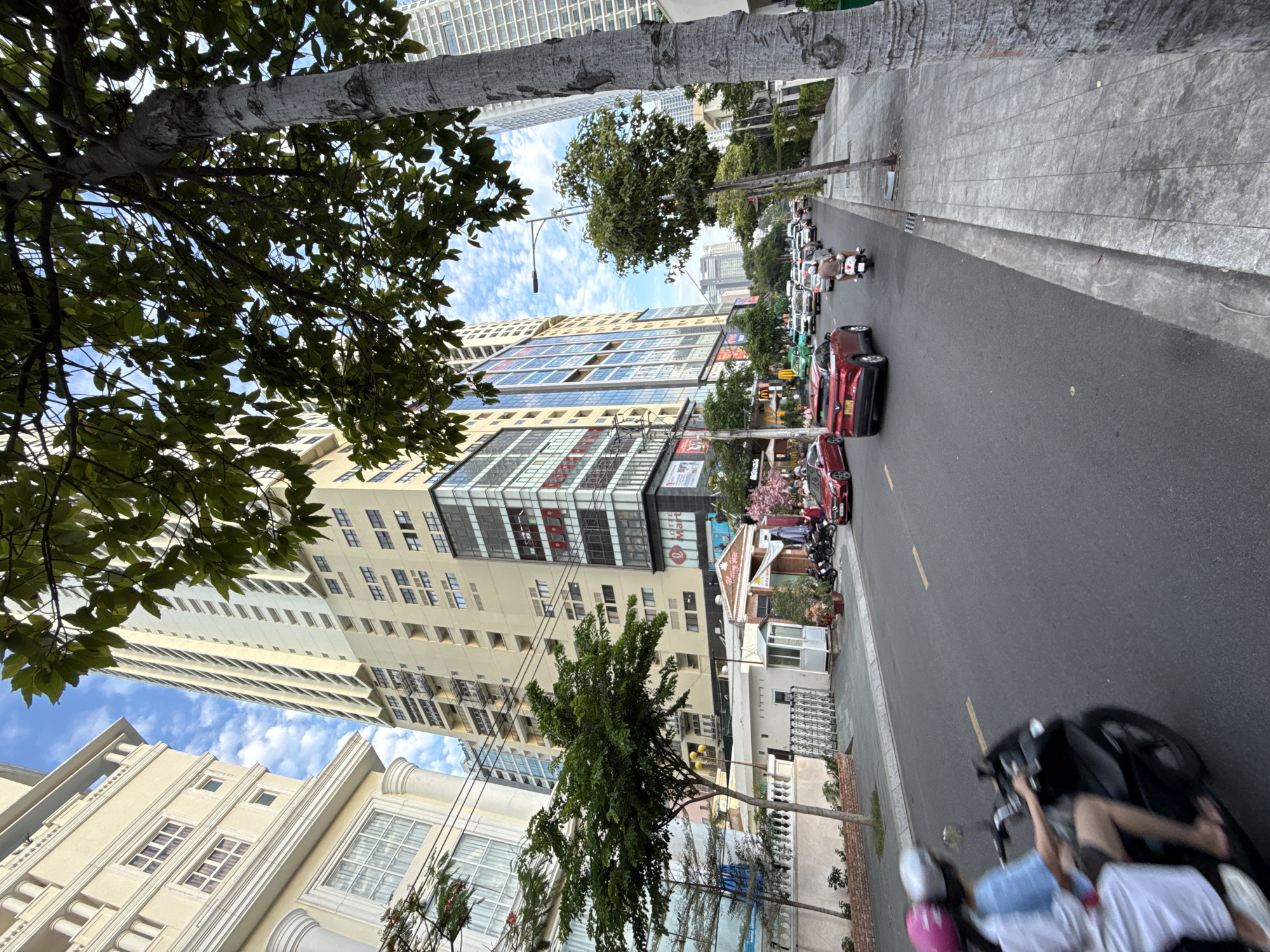 This is a street-level, low-angle shot taken on a clear day, capturing a bustling urban scene in Nha Trang, Vietnam. The foreground features a paved road with a concrete curb and sidewalk. On the road, a black motorcycle with two occupants, a driver and a passenger, is in motion, with the passenger wearing a white outfit and the driver in a blue top. The passenger is looking to the right, and both are wearing helmets. To the right of the road, trees with textured bark are visible along the sidewalk.
Further down the street, a dense traffic flow of cars and motorcycles is apparent. Several red cars, a green car, and various motorcycles are present. The buildings lining the street are tall and modern. On the left, a cream-colored building with classical architectural elements like columns and archways is visible, featuring multiple windows. Adjacent to it is a very tall, light-colored high-rise building with many windows and visible external staircases or fire escapes. On the right side of the street, another modern high-rise building, predominantly glass, is seen. Several commercial signs are visible on the buildings, including "LOTTE MART".
Trees with green foliage are scattered along the street, providing some shade. The sky above is blue with scattered white clouds, indicating fair weather. The overall impression is of a busy commercial district within a Vietnamese city.