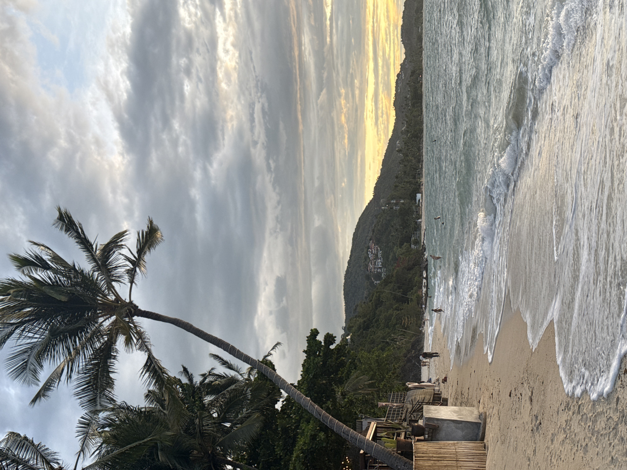 The image depicts a beach scene in Baan Tai, Thailand. The sky is filled with clouds, some tinged with the yellow light of the setting sun. A dark, densely vegetated hill rises along the coastline, with a few buildings visible on its slope. Waves break on the sandy beach, with people visible in the water and along the shoreline. In the foreground, several palm trees lean toward the right side of the image. A wooden platform or structure is also visible near the beach.