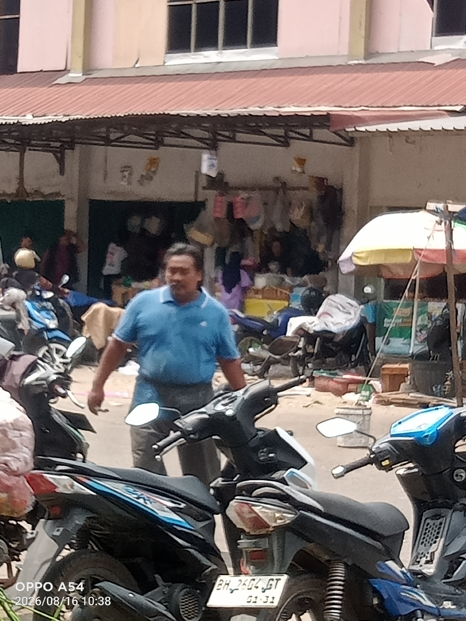 A middle-aged Indonesian man, wearing a blue polo shirt and dark pants, walks past several parked motorcycles in a busy outdoor market. He appears to be holding a cigarette. The market has a covered roof supported by pillars, and stalls are visible in the background displaying various items. The motorcycles, some of which have luggage on them, are parked on a paved surface. The overall scene suggests a bustling commercial area within the city of Jambi, Indonesia.