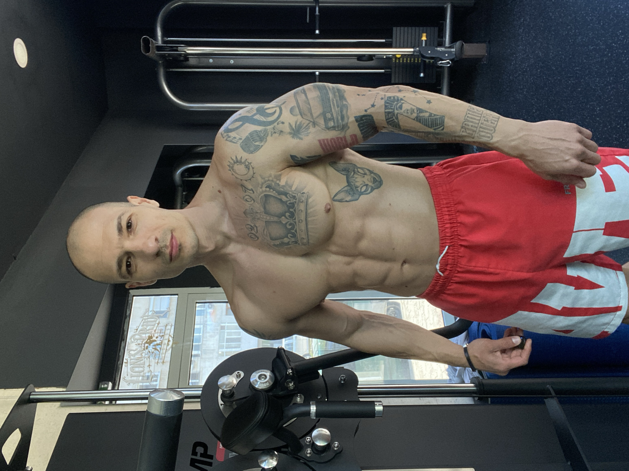 A muscular man stands in a gym. He is bald with tan skin and visible tattoos on his arms and chest. He wears red and white shorts. He is positioned in front of a workout machine. Through the gym window, buildings of Pleven, Bulgaria are visible.