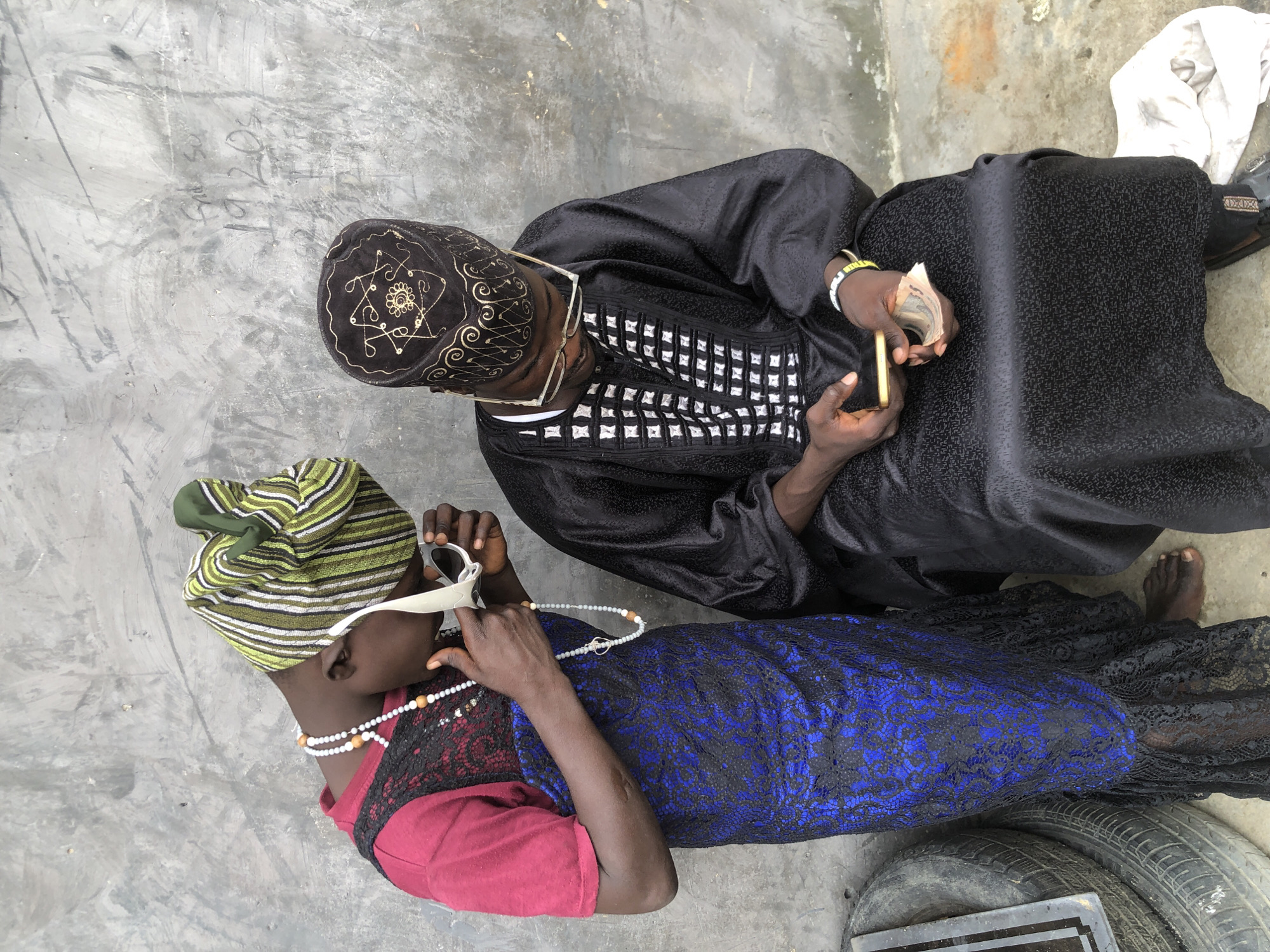 In a dusty outdoor environment in Ado, Nigeria, a person wearing a red shirt, black lace overlay, and a vibrant blue and black patterned dress stands to the left. They are adorned with a beaded necklace and are holding white sunglasses to their face. To the right, a person dressed in a traditional black garment and matching cap with gold embroidery is seated. They are looking down at a smartphone in their hands, which also hold a wad of cash. The backdrop is a concrete wall with visible graffiti. To the far left, a stack of tires and a metallic object are present.