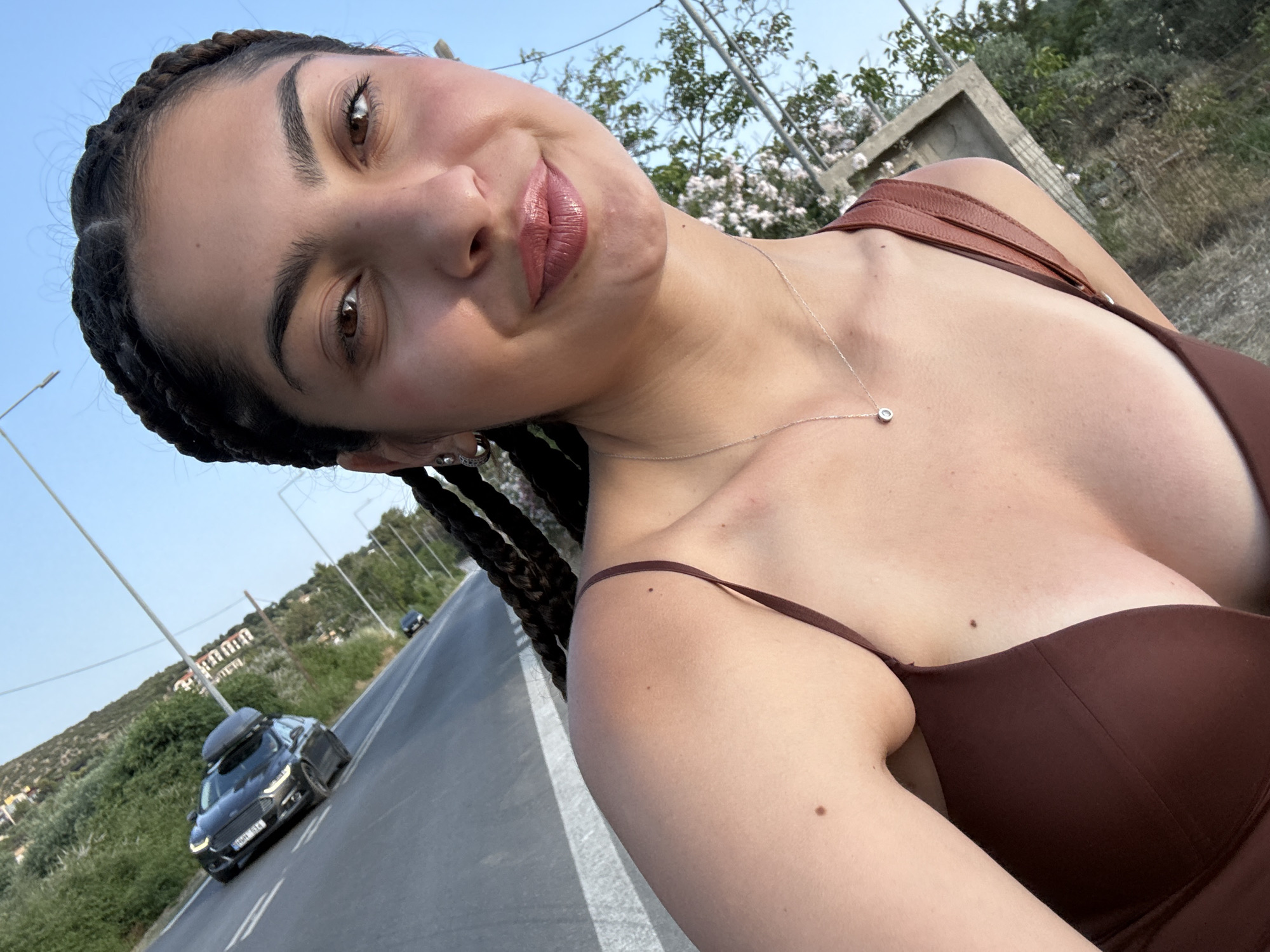 A woman with dark braided hair and brown eyes smiles at the camera in a selfie. She is wearing a brown bikini top and a silver necklace with a small pendant. She is standing on a road with a black car in the background driving away from the camera. The road is lined with green bushes and trees, and there are buildings on a hill in the distance. The sky is clear and blue. The location appears to be in Potos, Greece, as indicated by the surrounding landscape and the presence of a car with a license plate that might belong to the region.