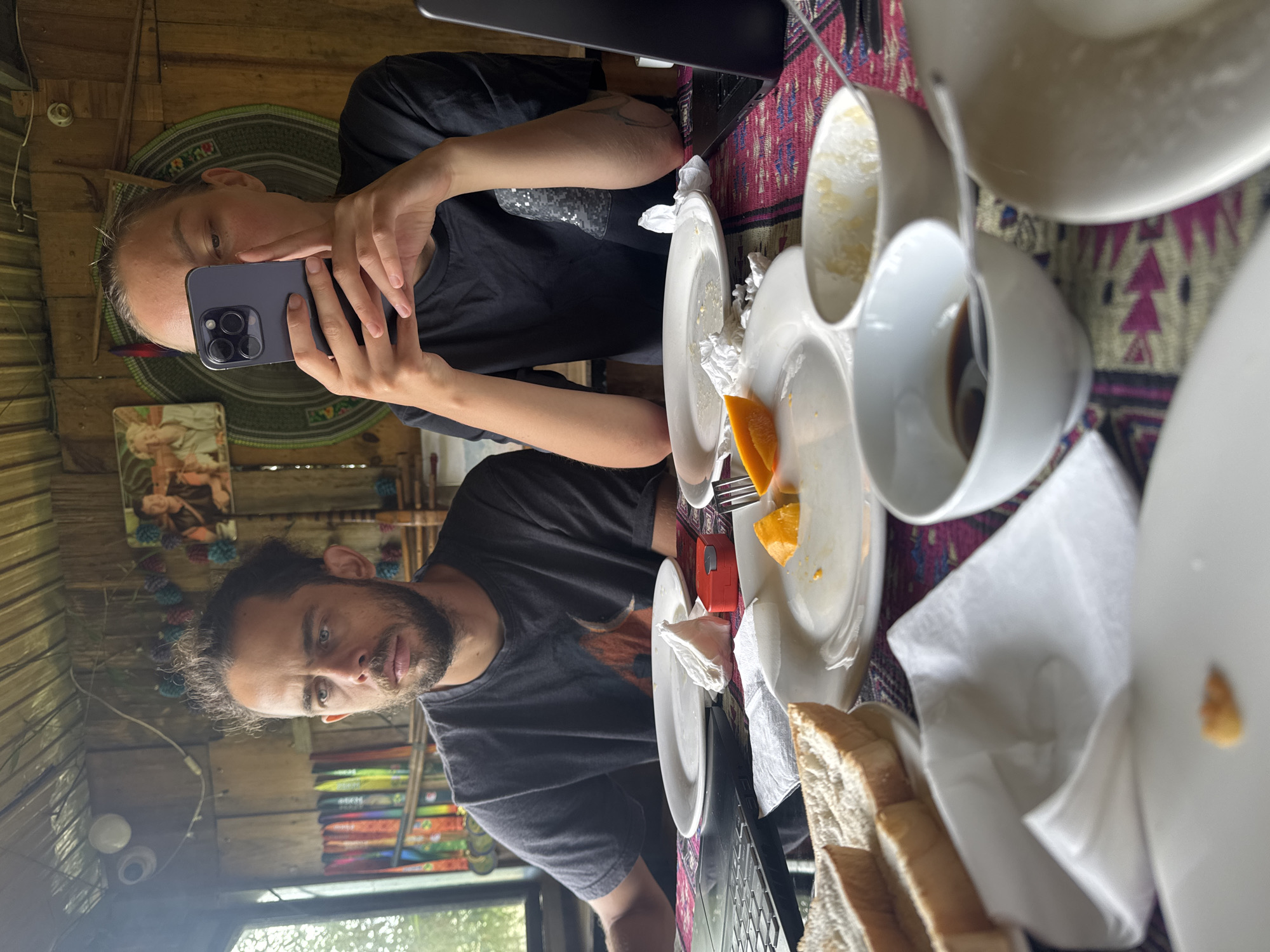 The image depicts two individuals, presumably a couple, at a table seemingly having breakfast or brunch in a rustic setting. One individual, seen from the chest up, is holding a smartphone, possibly taking a picture or video of the scene. The other individual is visible in the lower part of the frame, looking towards the camera. Both are wearing dark-colored shirts.
The table is covered with a patterned cloth and laden with remnants of a meal. There are several plates, some with leftover food like orange slices and crumbs, white napkins, a stack of bread slices, and cups, some appearing to contain coffee or another dark beverage.
The environment appears to be a wooden structure, potentially a cabin or a similarly constructed dwelling. The lighting suggests it is daytime, with natural light streaming through what appears to be a window. The setting has a slightly unkempt or lived-in quality.
There is no way to confirm the location in the city of Ngã Ba Kim, Vietnam.