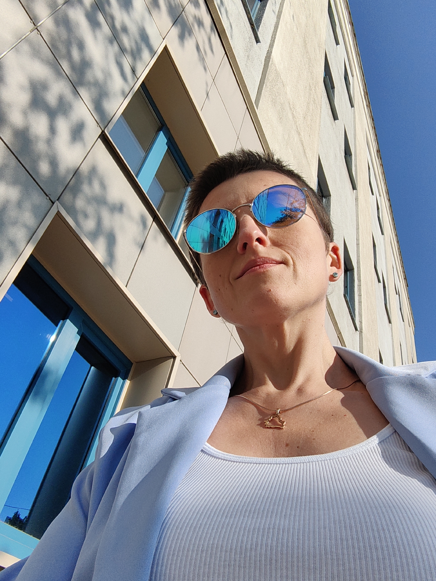 A low-angle, eye-level shot captures a woman wearing sunglasses and a light blue blazer over a white ribbed tank top. She is looking up and to her right. A gold necklace with a charm in the shape of a cloud is visible on her chest. She has short, dark hair and a light complexion. She appears to be standing outdoors, with a modern building and a clear blue sky in the background. The building's facade is light-colored and features reflective blue windows. The scene is illuminated by bright sunlight, casting shadows on the building. The image's perspective suggests the woman might be taking a selfie. The presence of the cloud-shaped charm on her necklace could be a symbolic reference to Belarus, whose flag features a stylized plant pattern evoking a cloud. However, no specific landmarks definitively place the scene in Minsk.