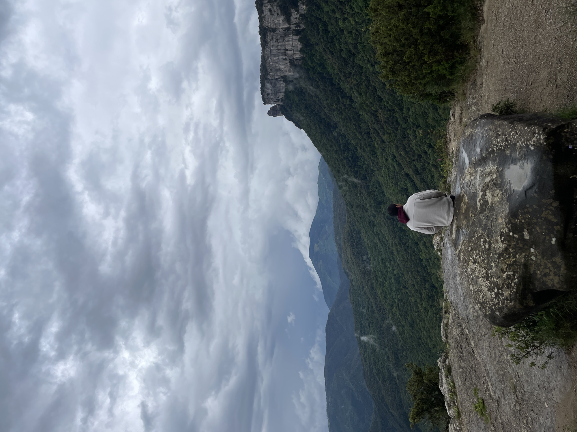 The image depicts a person sitting on a large, lichen-covered rock outcrop at a high altitude. The person is facing away from the camera and wears a light-colored top and what appears to be a scarf or garment with a reddish hue near the neck. The background features a dramatic landscape with a steep, heavily forested mountain or cliff face to the right. A complex of mountains fades into the distance, obscured by a sky filled with heavy, overcast clouds. The scene conveys a sense of altitude and the vastness of the natural environment. Given the mention of Rupit i Pruit, Spain, it is possible the image was taken within or near the natural areas of that region.