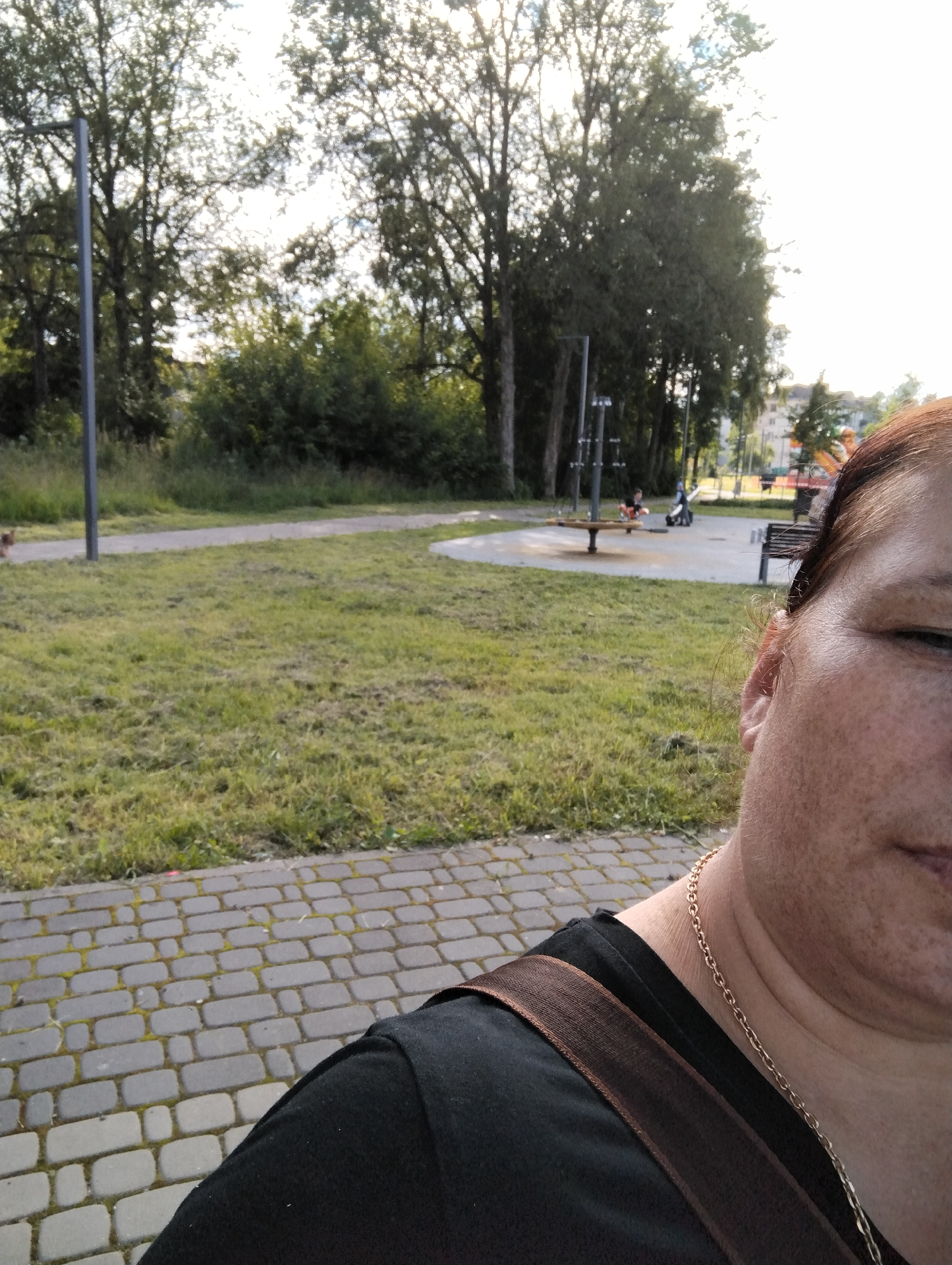 The image is a selfie taken outdoors, likely in a park or recreational area in the city of Kingisepp, Russia. In the foreground, a woman's face and upper body are visible on the right side. She has brown hair, fair skin with freckles, and is wearing a black t-shirt with a brown strap across her shoulder and a gold chain necklace. The background shows a paved walkway, a grassy area, trees, and a playground with swings and other equipment. Several children are visible in the playground. The lighting suggests it is daytime, possibly late afternoon, with the sun casting a bright light in the sky.