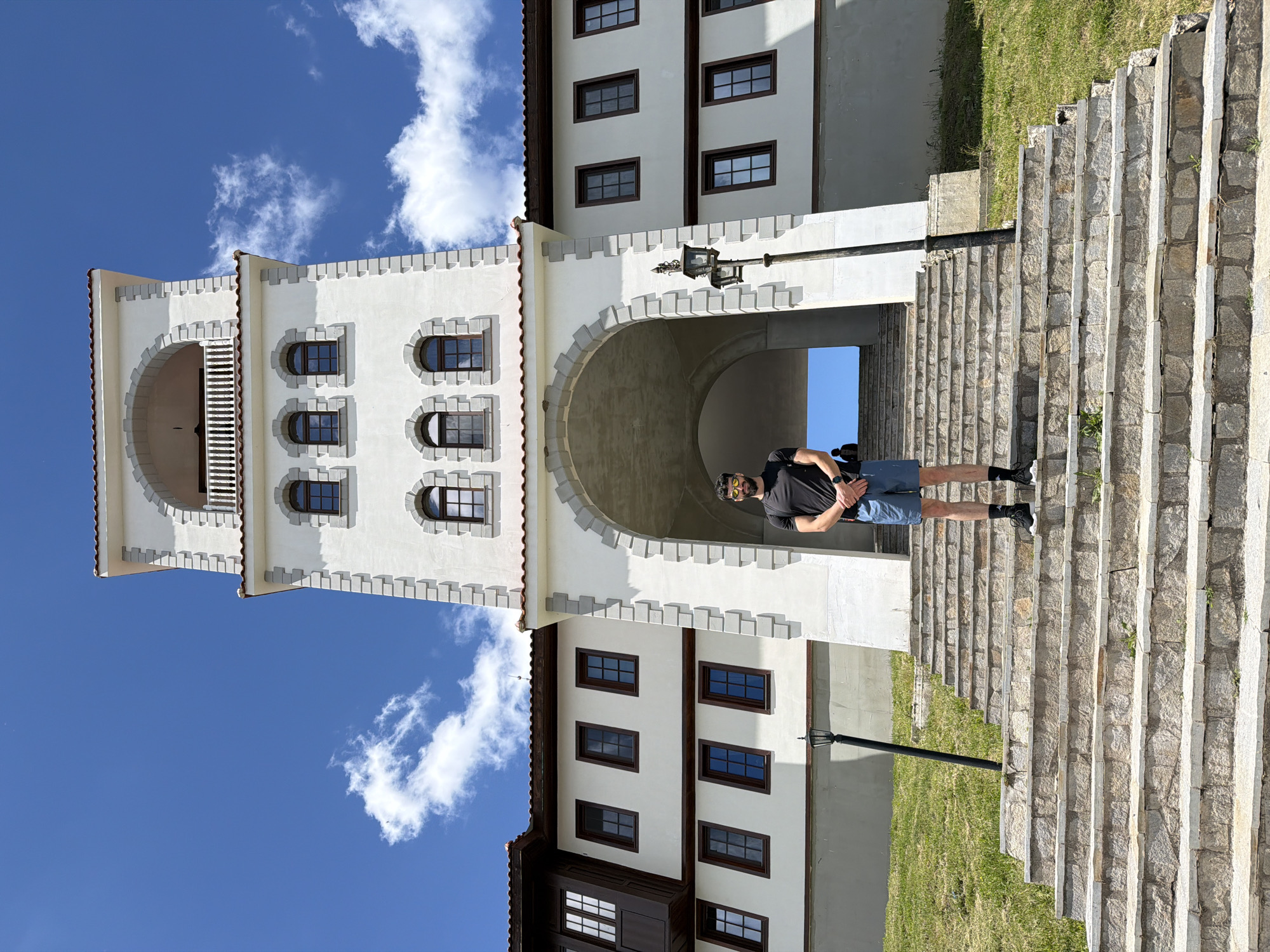 A man stands on a stone staircase in front of a white, multi-story building with a prominent tower. The building features arched doorways, multiple windows, and a red tiled roof. The sky is blue with scattered white clouds. The staircase is flanked by green grass.