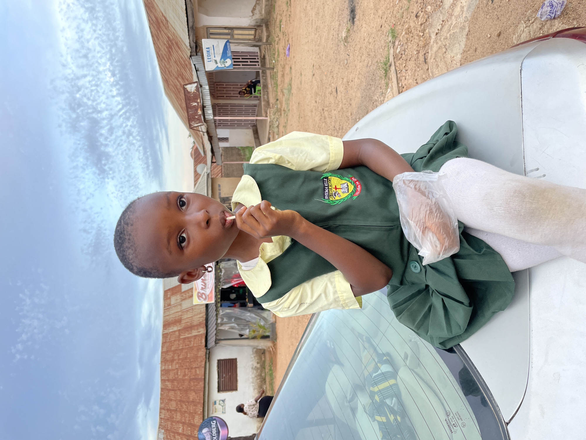 A young girl in a green and yellow school uniform is sitting on the hood of a silver car. She has a lollipop in her mouth. The school uniform bears the inscription "RHEMAVILLE ACADEMY." She has white socks on and a clear plastic bag around her lower leg. The background shows a street scene in what appears to be Jos, Nigeria. There are shops and buildings with corrugated iron roofs. A Goldberg sign is visible in the background. Other pedestrians can be seen in the street. The sky is overcast.