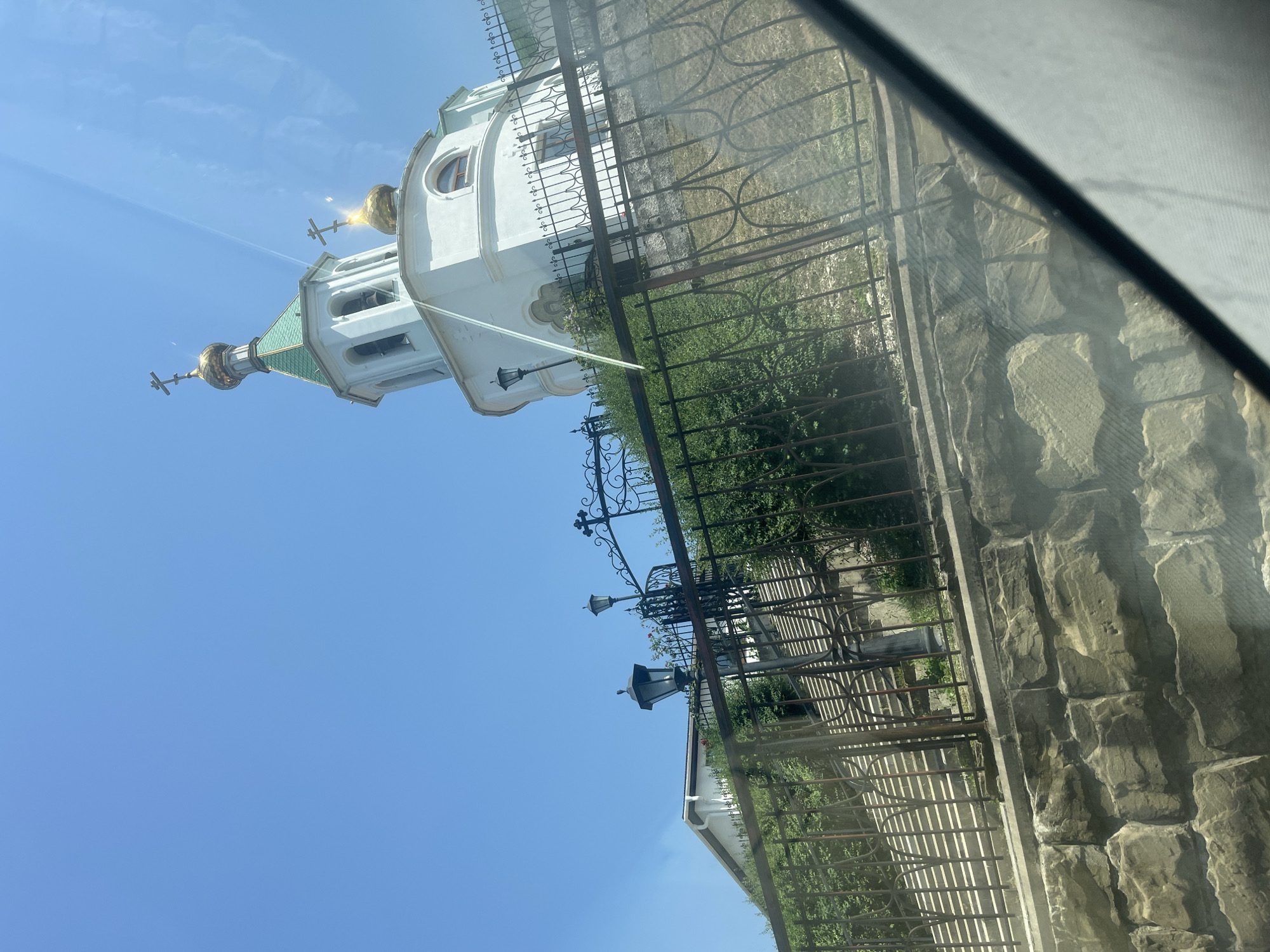 A white church with a green roof and gold domes stands on a hill against a clear blue sky. A wrought-iron fence with decorative elements runs along the foreground, separating the viewer from a stone wall and a grassy slope. Steps ascend the hill towards the church, flanked by green bushes and illuminated by ornamental street lamps. The image appears to be taken from inside a vehicle, with a reflection on the glass.