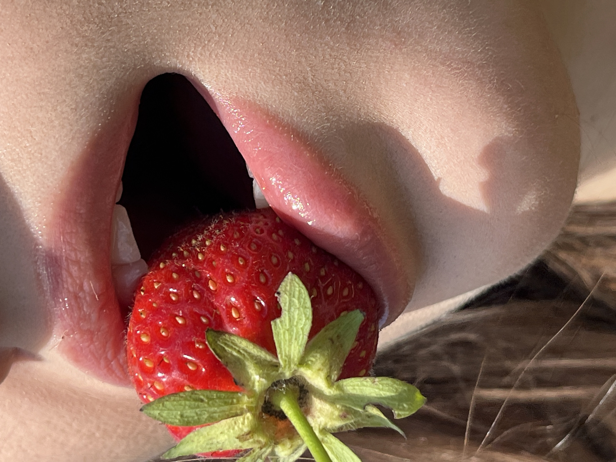 A close-up, high-angle shot shows a person's mouth and chin holding a ripe strawberry. The strawberry is red with visible seeds and green leafy stems. The person's lips are slightly parted, revealing a glimpse of their teeth and tongue. The skin on the chin and cheek is visible, with a shadow cast across the lower right side of the face. The background is blurred, showing dark strands that appear to be hair. The image is taken outdoors, suggested by the natural lighting.
