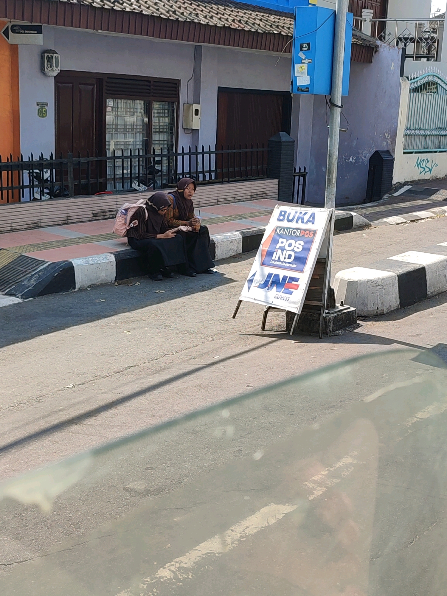 The image captures a street scene in Magelang, Indonesia, on a sunny day. In the foreground, a white A-frame sign displays logos and text, advertising "BUKA KANTORPOS POS IND Logistic Indonesia" and "JNE EXPRESS". Two young women, dressed in hijabs and traditional Indonesian attire, are seated on a concrete curb in front of a building. One woman, on the right, holds a takeaway cup and appears to be speaking, while the other, on the left, looks down at her phone, with a pink backpack beside her. Behind them, the building features a dark brown wooden door and windows with visible interior curtains. A black metal fence runs along the front of the property. To the right of the building, a blue utility box is mounted on a pole, adorned with several stickers. The street itself is paved asphalt, with a distinct crosswalk marked by white and black striped concrete barriers. Graffiti, a stylized "MSS Y", is visible on the wall to the right. The overall setting suggests a commercial or residential area within the city.
