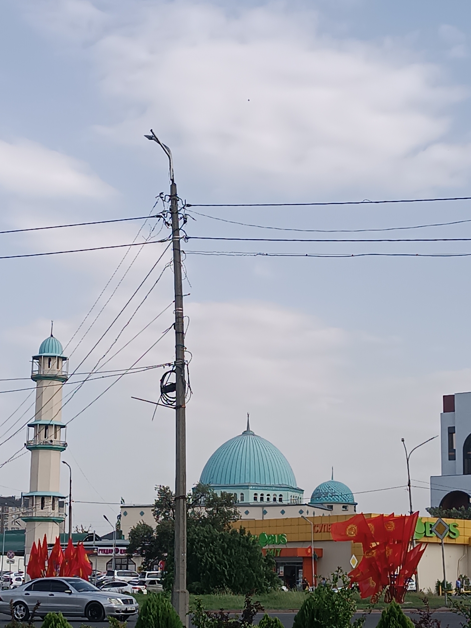 In Bishkek, Kyrgyzstan, a scene unfolds with architectural and civic elements under a pale, cloudy sky. Dominating the background are two domed structures, likely part of a mosque complex, with a prominent light blue dome and a smaller, matching dome. To the left, a slender minaret with a light blue cap stands tall. In front of these structures, a commercial building with a yellow facade and signage is visible, featuring the Cyrillic word "СИЛЕ" and partially obscured text that might spell out "GLOBUS." A silver sedan is parked on the street in the foreground. A utility pole with a network of wires stretches across the frame, a common sight in urban environments. Red flags with what appear to be national symbols are clustered near the street, suggesting a cultural or national event. The environment is moderately green with trees and bushes adding to the urban landscape.