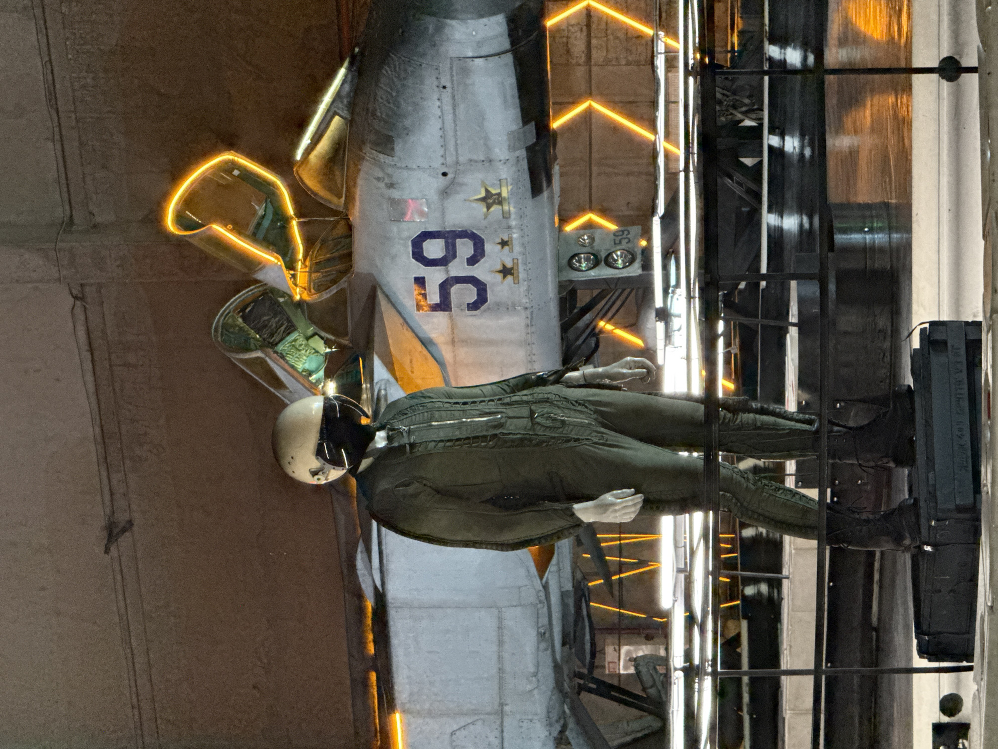 The image depicts a mannequin dressed in a green flight suit and helmet, standing in front of a fighter jet. The jet has the number "59" painted on its side, along with two stars. The cockpit canopy of the jet is open and illuminated by orange neon lights, which also highlight the exterior of the aircraft and the surrounding area. The scene appears to be indoors, possibly in a museum or exhibition space, with concrete walls in the background. The floor is dark and reflective, mirroring the neon lights. The lighting creates a dramatic and modern atmosphere, contrasting with the vintage appearance of the aircraft and flight suit. This scene is located at the Central Naval Museum in Saint Petersburg, Russia, which showcases a collection of naval artifacts, including aircraft.