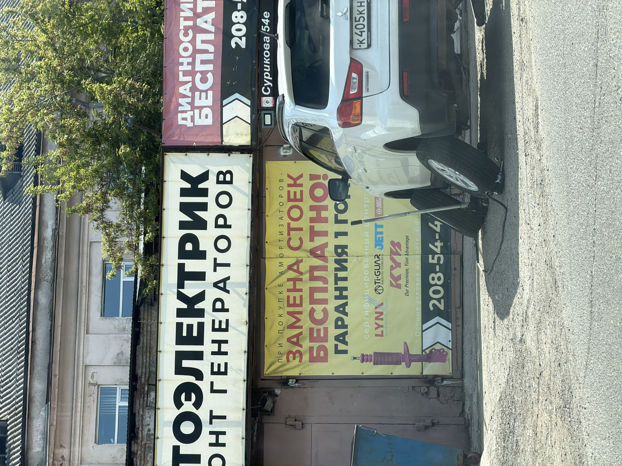 The image depicts a street scene in Krasnoyarsk, Russia, with parked vehicles and building facades covered in advertisements. A white Mitsubishi car, registration number K405KH, is parked near a building with various signs. These signs promote services such as generator repair ("ТОЭЛЕКТРИК ОНТ ГЕНЕРАТОРОВ"), free strut replacement with amortizer purchase ("ПРИ ПОКУПКЕ АМОРТИЗАТОРОВ - ЗАМЕНА СТОЕК БЕСПЛАТНО! ГАРАНТИЯ 1 ГО"), and a diagnostic service ("ДИАГНОСТИК БЕСПЛАТ"). Brands like KYB, Lynx, Tigar, and Jette are advertised. A phone number (208-54-4) is prominently displayed. The location is identified as "Сурикова 54е." A spare tire and car jack are also visible near the parked car. The overall impression is of an automotive repair shop or service center.