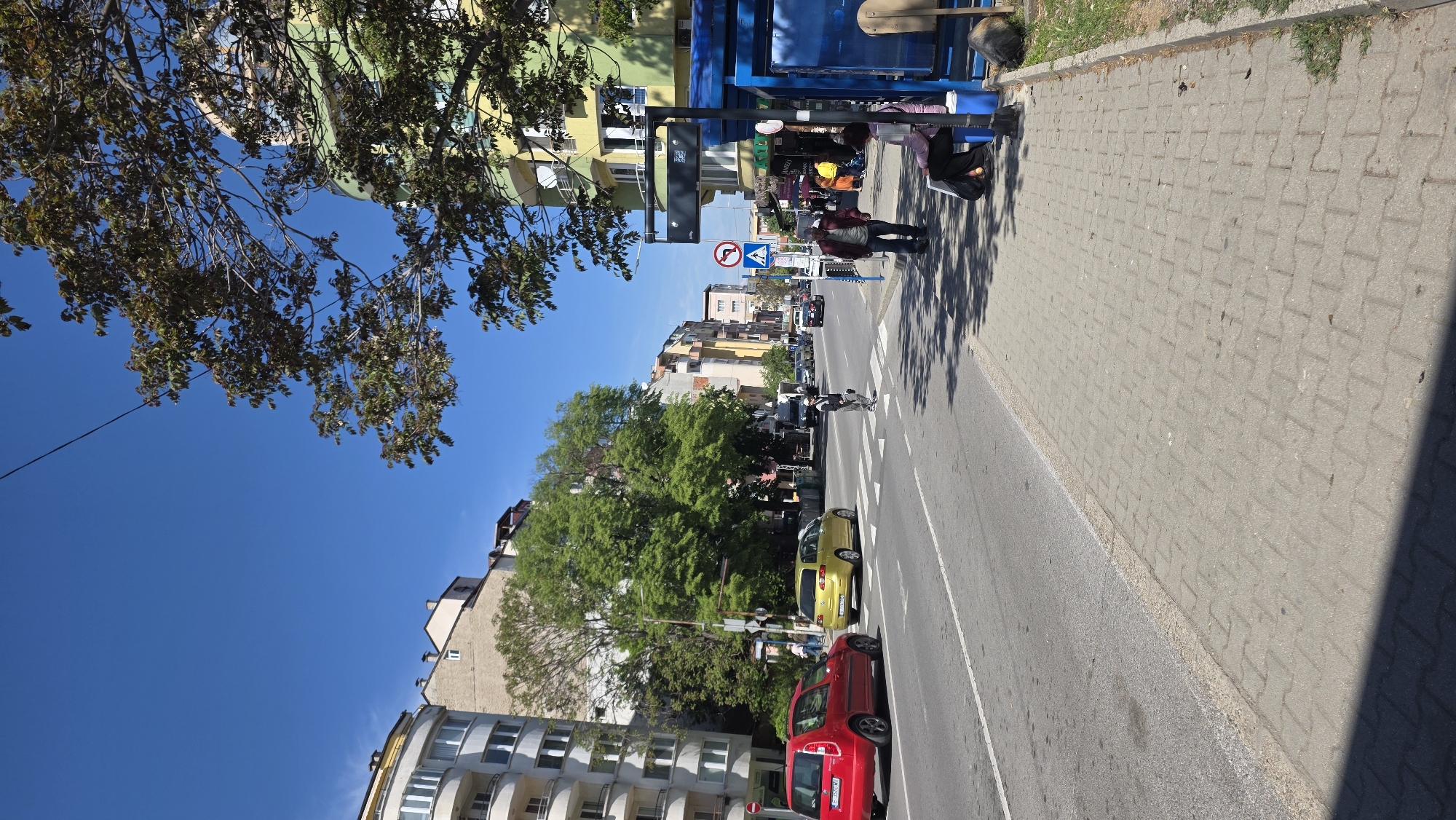The image shows a street scene in Blagoevgrad, Bulgaria. The view is along an urban street with buildings of varying heights on either side. A red car and a yellow car are parked on the right side of the road. Pedestrians are present on the sidewalk to the right, waiting at a bus stop. One person is crossing the street on a pedestrian crossing. Traffic signs are visible near the bus stop. The sky is blue and clear. Branches from trees are visible on the left, adding a touch of nature to the scene.