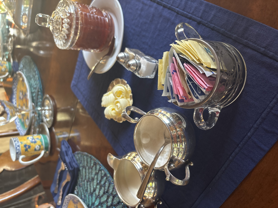 A meticulously arranged breakfast or dining table setting is depicted indoors, presumably in the city of Monterey, United States of America. The scene features a polished wooden table, partially covered by a dark blue fabric placemat or runner, and a teal-patterned circular placemat to the left.
In the foreground and midground, a collection of condiments and serving items is displayed on the blue placemat. On the far left, a silver creamer pitcher contains a white liquid. Adjacent to it, a silver sugar bowl holds white granulated sugar and a small silver spoon. To the right of these, a small scalloped silver dish presents three carefully shaped butter curls. A clear glass salt and pepper shaker set stands next to the butter dish. Further to the right, a clear glass jar with a decorative lid, containing a reddish-brown preserve (likely jam), rests on a small white saucer with a silver spoon. In the bottom right, a clear glass cup-shaped holder is filled with an assortment of individually wrapped sugar substitute packets in yellow, pink, and blue colors.
In the blurred background, parts of a place setting are visible, including a mug with yellow and blue floral patterns, a decorative plate with similar motifs, a clear wine glass, and silver cutlery (fork and knife) resting on a folded dark blue napkin. No individuals are present in the frame; the items are prepared for consumption.
