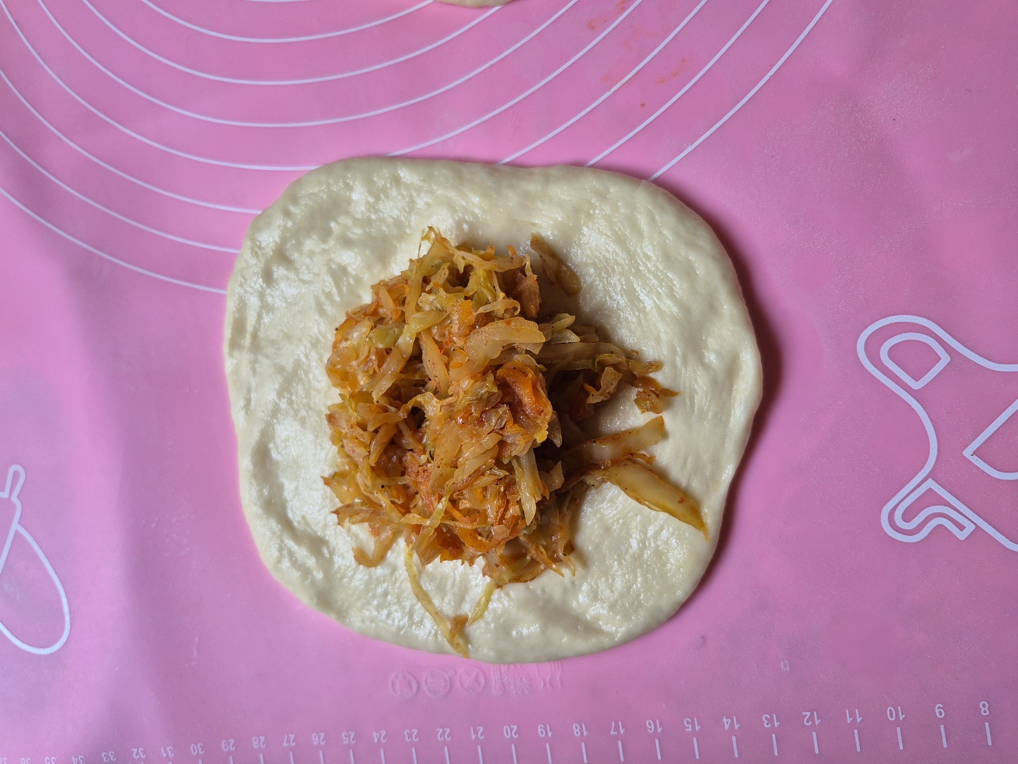 A top-down, close-up shot shows a flat piece of raw dough on a pink silicone baking mat. The dough is roughly rectangular with rounded corners and has a filling of shredded vegetables, likely cabbage and carrots, piled in the center. The vegetables appear to be cooked and seasoned, with a reddish-orange hue from spices. The baking mat has concentric white circles and a ruler marked in centimeters along the left edge, with numbers from 8 to 37 visible. The background is a solid pink surface. The scene appears to be indoors, in a kitchen or food preparation area. No people are visible in the image.
