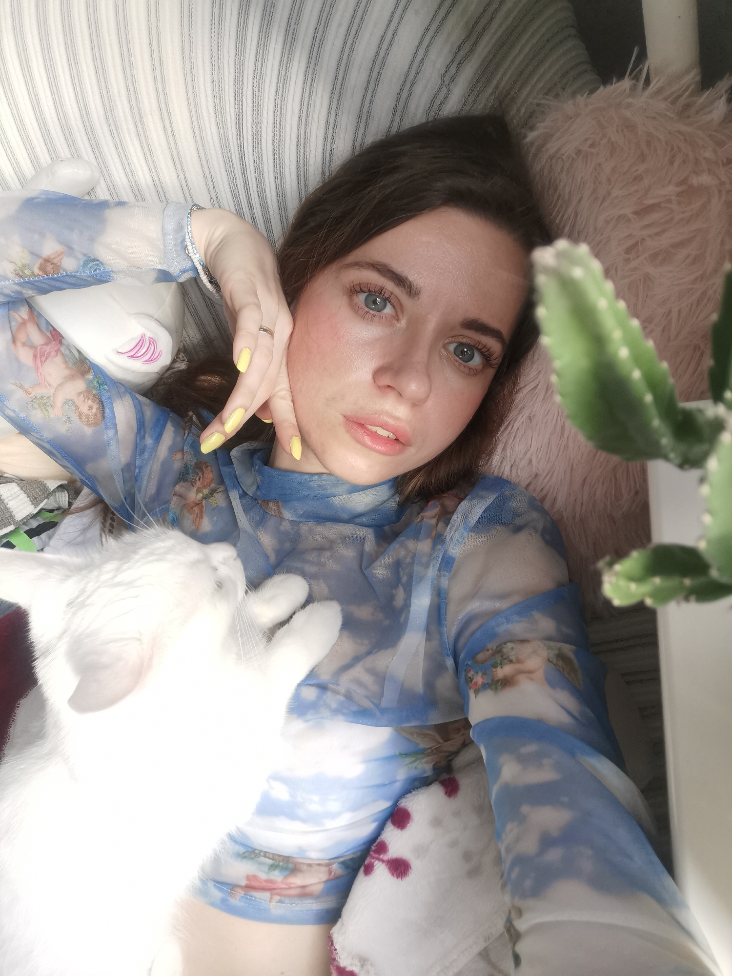 A woman lies on a striped cushion with a white cat. She wears a long-sleeved sheer shirt with a blue and white cloud design. A green cactus stands next to her. Her nails are painted yellow. The location appears to be indoors. The city and specific location are unknown.