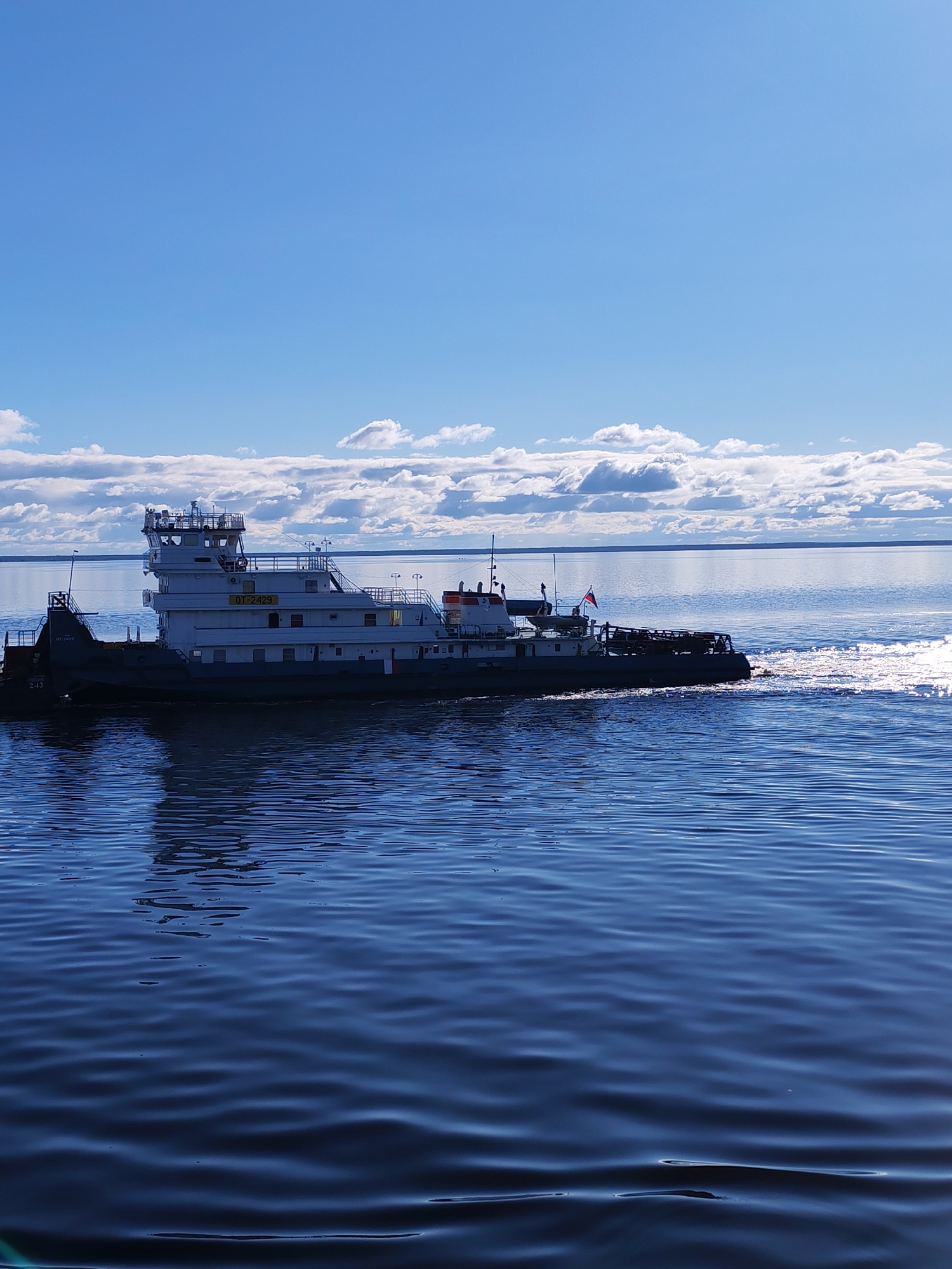 A tugboat is traveling on the waters of what appears to be a large body of water under a partially cloudy sky. The tugboat has a white upper structure and a darker hull. The Russian flag is visible on the rear of the boat. Sunlight reflects on the water's surface, creating a shimmering effect. The location is Mikhalevo, Russia.