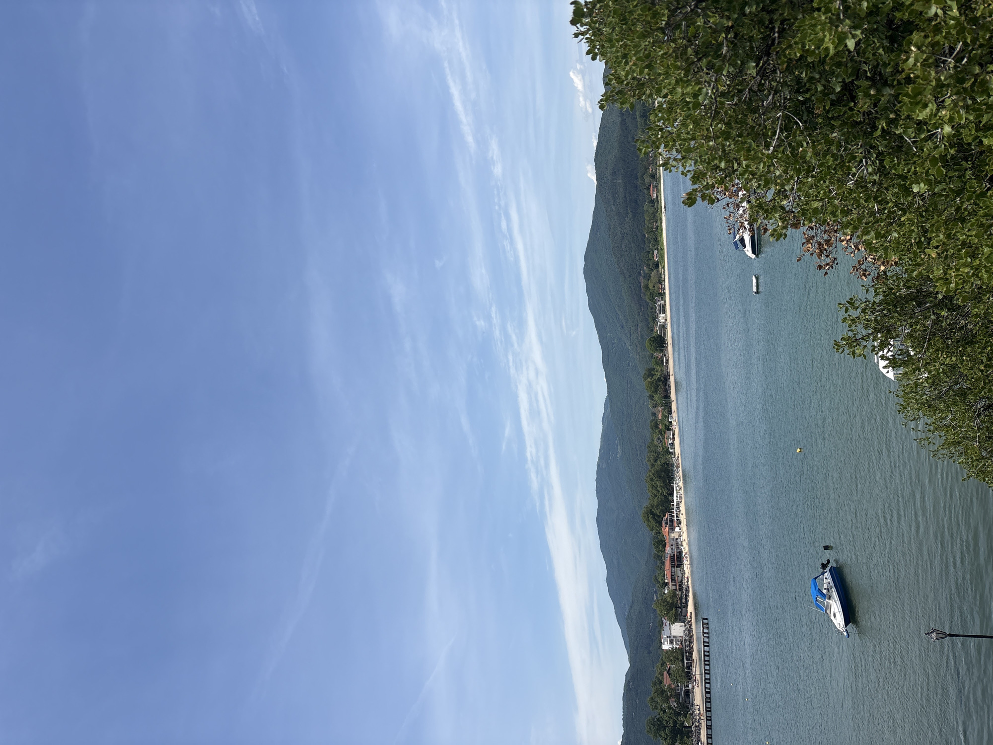 The image shows a coastal scene in Olympiada, Greece, with a view of the Aegean Sea. In the foreground, lush green foliage partially obscures the view of the water. The sea is calm, with several boats visible. A small white and blue motorboat is in the center of the frame, with a black object, possibly an anchor or a buoy, next to it. Further out in the water, another larger white boat is partially visible near the shore.
In the middle ground, a long wooden pier extends into the sea from a populated coastline. Along the shore, there are buildings with red-tiled roofs, characteristic of Greek architecture, interspersed with trees and a sandy beach dotted with sunbathers and umbrellas. The beach appears to be a popular spot, with people relaxing and enjoying the seaside.
The background features a range of green, rolling hills that form a natural backdrop to the coastal town. The sky above is a clear, bright blue, with scattered wispy white clouds, indicating fair weather. The overall impression is of a serene and picturesque summer day in a Greek coastal resort.
