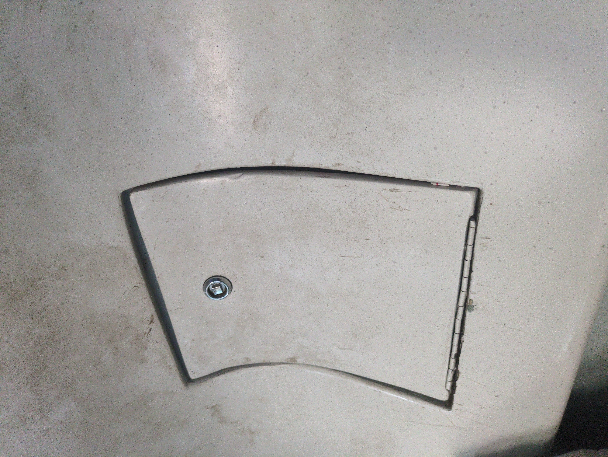 The image shows a light-colored, textured surface with a rectangular, inset panel. The panel has a curved outer edge and a flat inner edge where it meets the surrounding surface. A circular metal lock mechanism is visible on the upper portion of the inset panel. The surrounding surface appears weathered and has small, dark speckles scattered across it. The bottom edge of the inset panel shows a series of small hinges. The image does not provide any information to identify its location in the city of Aruki, India.
