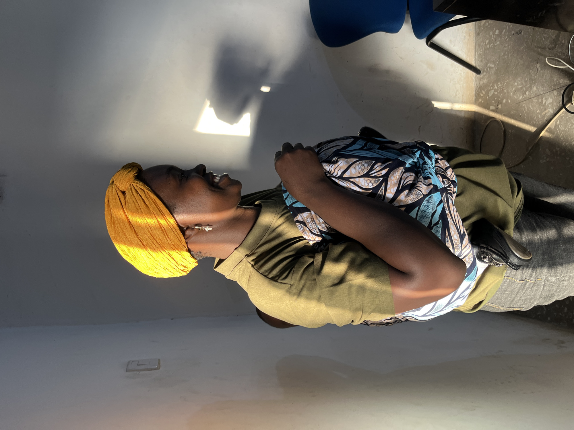 A low-angle, vertical shot captures a person wearing a bright yellow headwrap and an olive green shirt. The individual is facing to the left, with their head tilted back and mouth open in a laugh. They are holding a patterned fabric, possibly a scarf or shawl, adorned with blue and beige floral motifs, against their chest. The sunlight streams in from the top left of the frame, casting a bright glow on the wall and creating stark shadows. The person appears to be sitting in a room with a light-colored, possibly concrete floor. In the background, the leg of a dark blue chair and a black desk leg are visible. The scene suggests a casual, perhaps indoor setting during the day, with the strong sunlight indicating it is either morning or late afternoon. The overall mood is cheerful and relaxed. There is no visible text in the image.