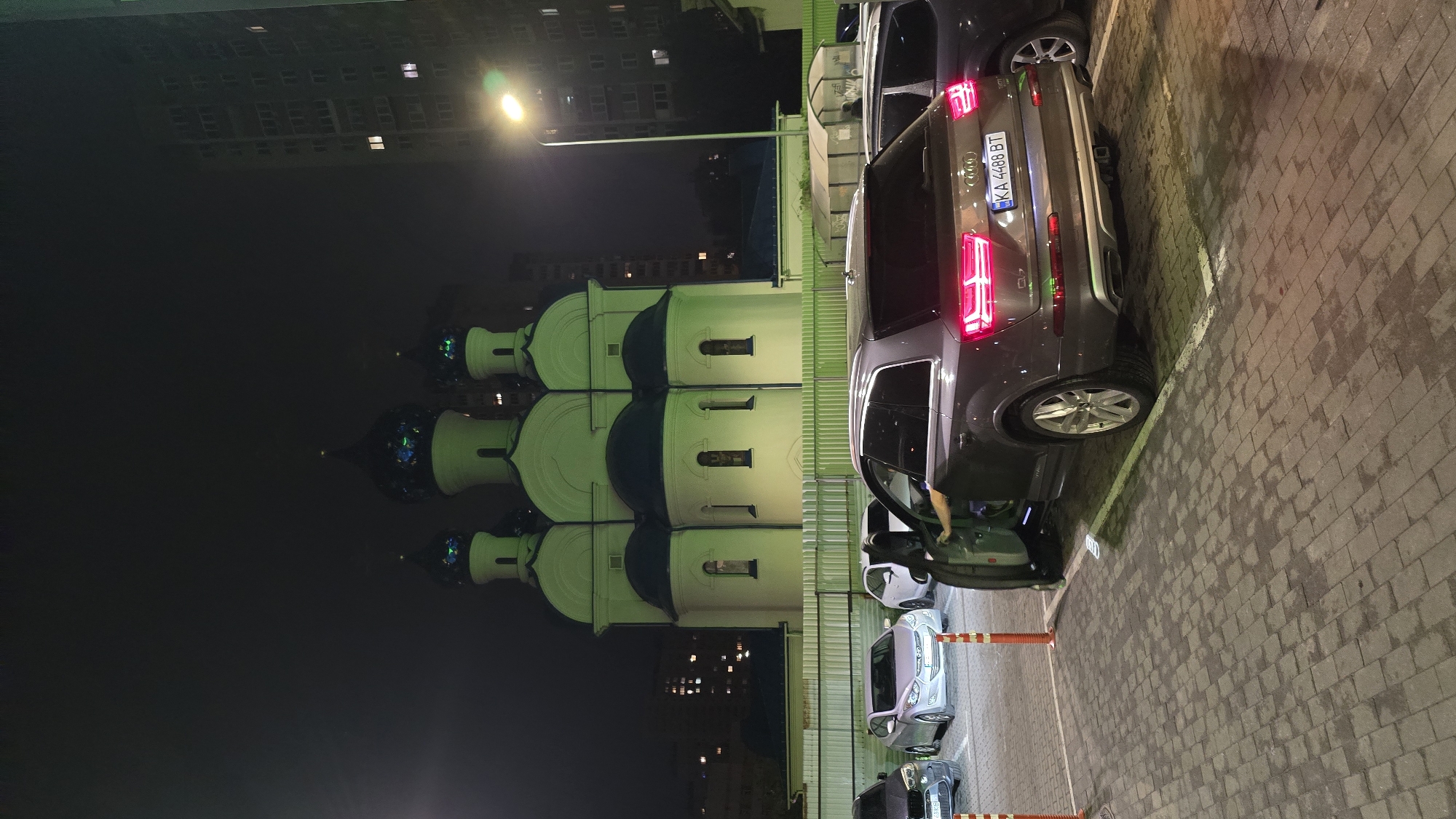 The provided image is rotated 90 degrees clockwise. When viewed upright, it depicts a nocturnal urban scene featuring a distinctive church-like building, several parked cars, and city infrastructure.
In the mid-ground, a large building resembling an Eastern Orthodox church dominates the left side, illuminated with a vibrant green light. It features multiple onion domes, some dark and others reflecting the green light, and white walls with small, recessed windows.
To the right, a parking area with brick-like pavers holds several vehicles. The most prominent is a dark gray or brown Audi Q7 SUV, seen from its rear-right side, with its red LED taillights glowing. A person's arm and leg are visible within the opened driver's door, indicating someone is either entering or exiting the vehicle. The Audi's license plate, "KA 4488 BT," identifies the location as the Kyiv region of Ukraine. Behind the Audi, a lighter-colored sedan and other parked cars are visible, separated by orange and white traffic bollards.
A tall street light with a bright, glowing fixture stands on the right, casting light onto the parking lot. In the background, taller city buildings with illuminated windows can be faintly discerned against the dark night sky. The scene captures a blend of modern urban life with traditional architecture under artificial night lighting.