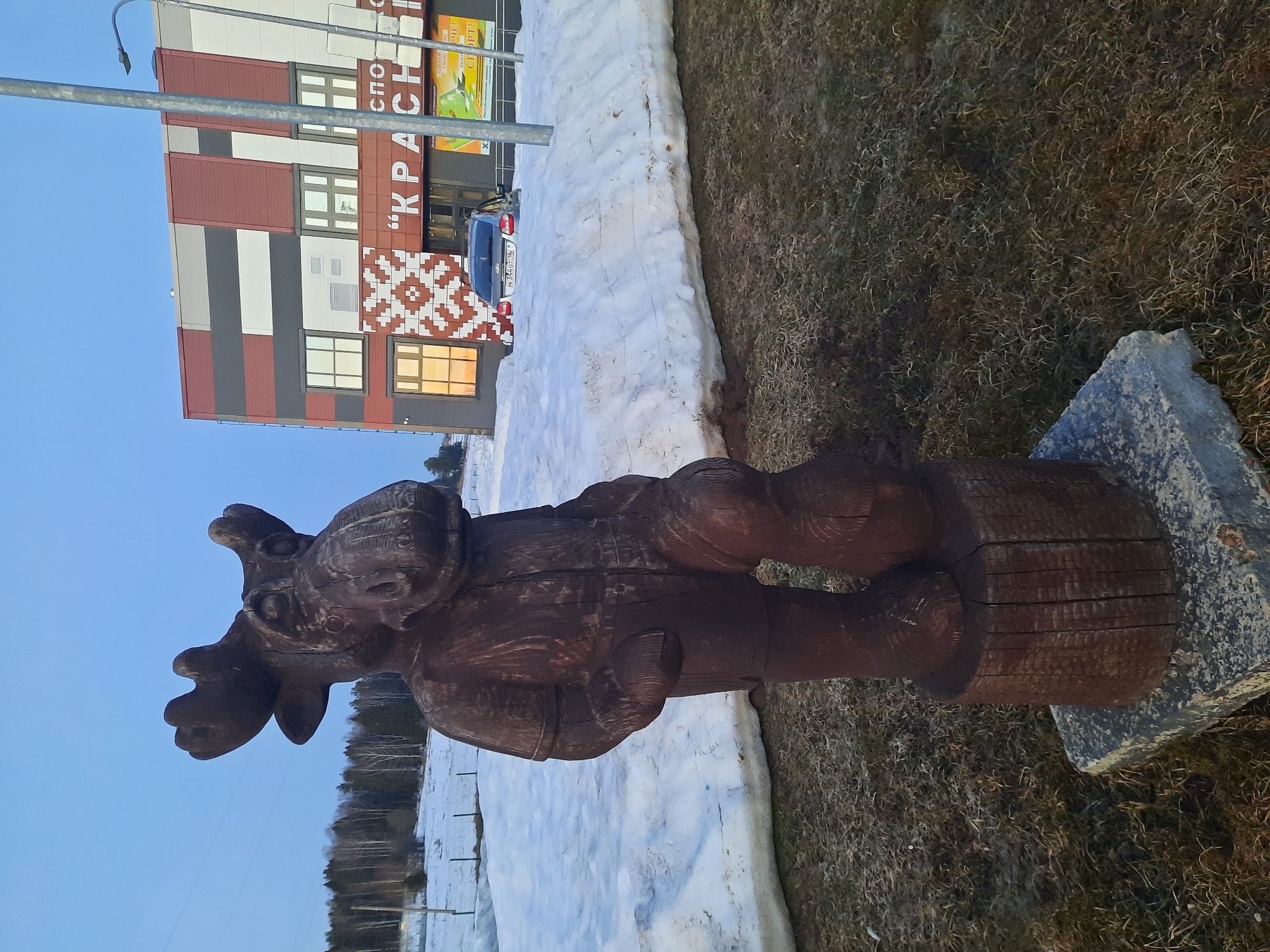 The image shows a wooden sculpture of a moose on a stone base in what appears to be a partially snow-covered area with grass. In the background is a modern building with red, white, and grey panels, bearing the inscription "KPACH" (possibly part of "KPACHO"). A streetlamp stands to the left of the building. Given the context that the location is Krasnogorskoye, Russia, it is likely a public art piece or a decorative element within the town. A dark-colored car is partially visible behind a snow bank in front of the building. The sky is blue, suggesting daytime. The environment seems to be a park or an open area with sparse trees in the far background.