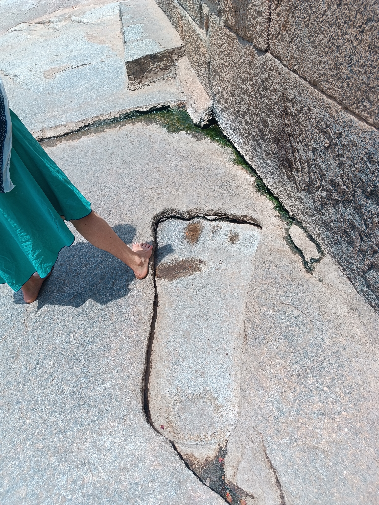 A full shot shows a person in a green skirt standing near a large, carved footprint in a stone surface. The stone surface appears to be part of a larger structure, possibly a building foundation or a pavement, given the presence of large stone blocks in the background. Green moss or algae grows in the crevices between the stones and along the base of the stone structure, indicating the presence of moisture. The person's right foot is positioned near the edge of the carved footprint, as if to compare the size. The scene is situated in the city of Unknown, Unknown.