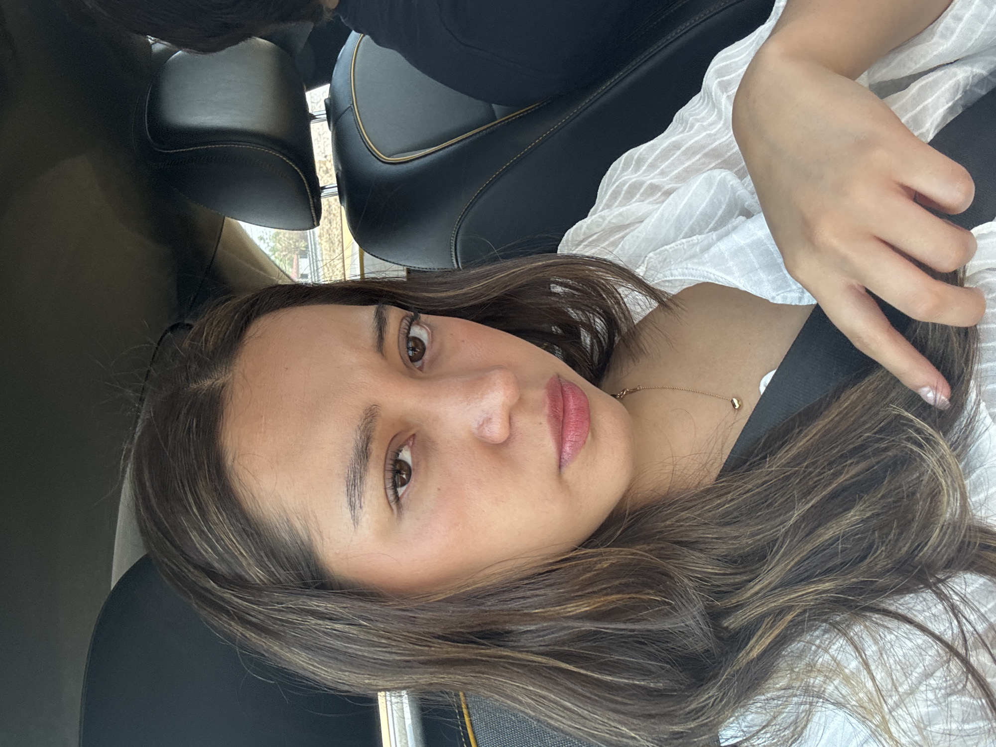 A woman with medium-length brown hair with blonde highlights is in the passenger seat of a car, wearing a white shirt. She is wearing a seatbelt. The interior of the car is black leather. The location is unknown and cannot be determined.