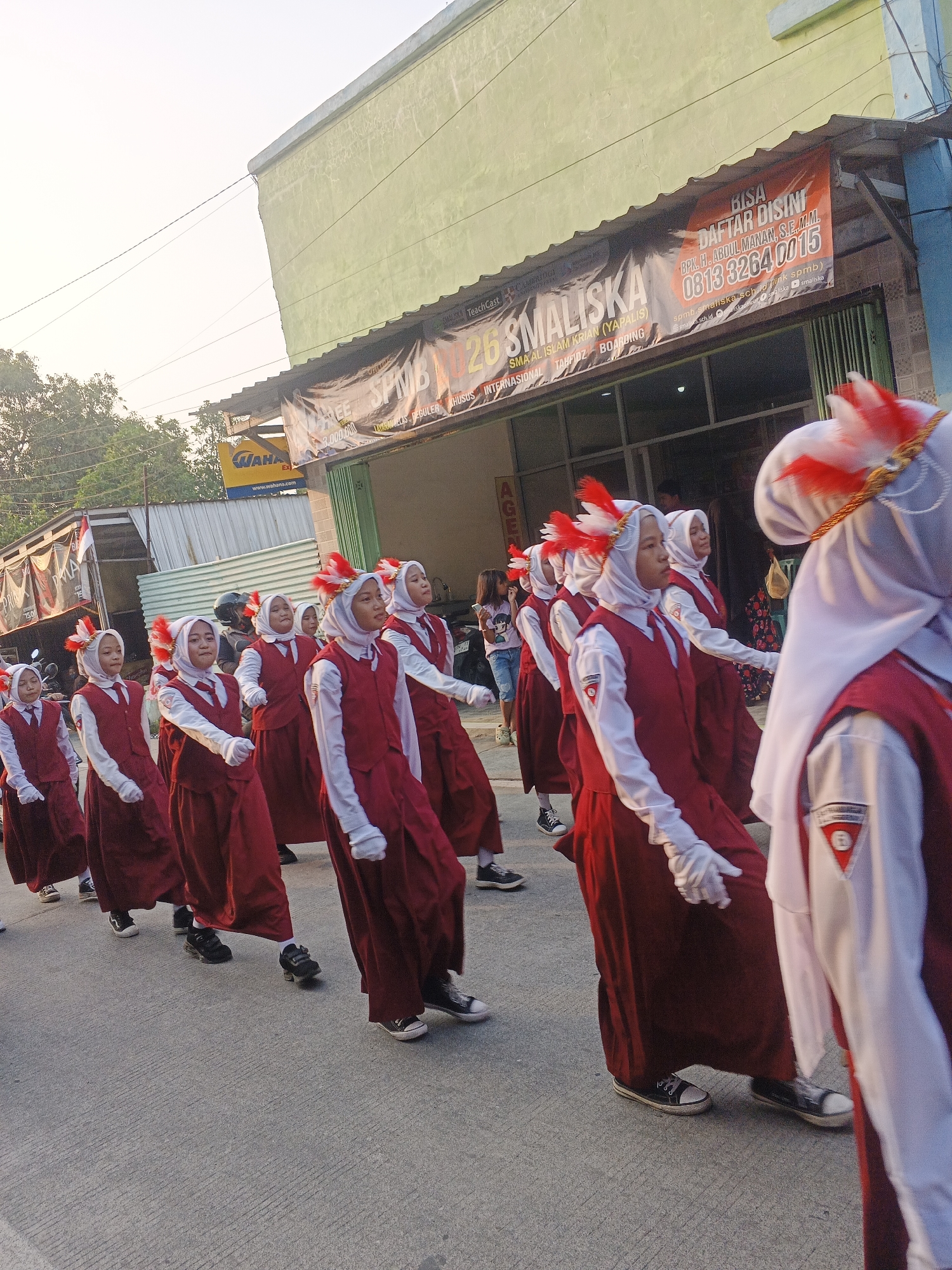 In Bogem Pinggir, Indonesia, a group of young girls in matching uniforms are participating in a parade. They are wearing white shirts with red vests and skirts, white gloves, and white headscarves adorned with red and white feathers. The girls are walking in a formation on a paved street, with buildings lining one side of the road. One building has a large banner with text that includes "BIISA DAFTAR DISINI" and a phone number. The other side of the street has trees and more buildings. The sun is shining, casting a warm light on the scene.