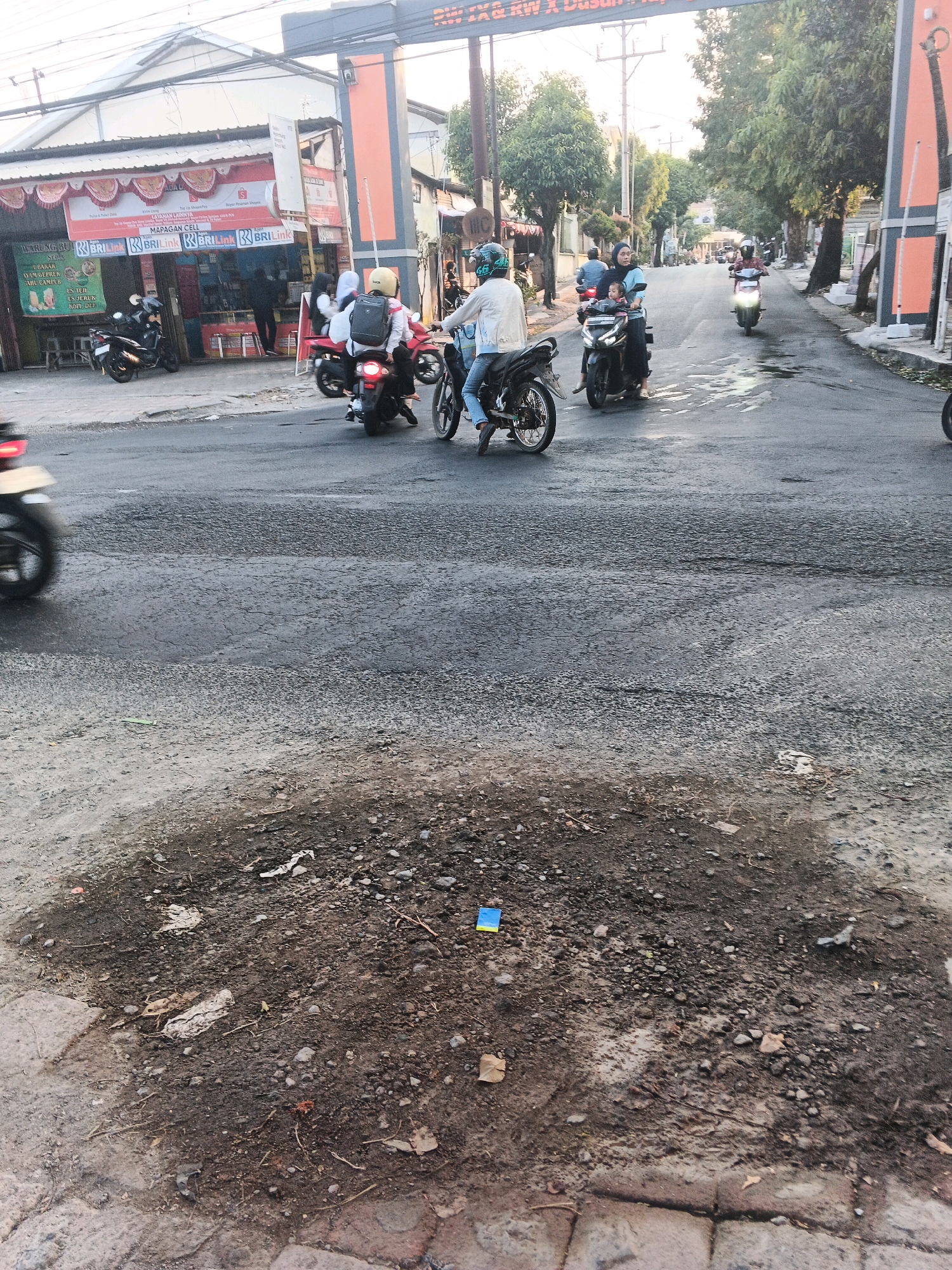 This image depicts a street scene in Ungaran, Indonesia, with an emphasis on the condition of the road and the presence of motorcycles. The foreground is dominated by a pothole in the asphalt, filled with loose gravel and dirt, with some scattered debris including a small blue and yellow object, possibly a piece of plastic. Beyond the pothole, the asphalt road surface appears cracked and uneven in several areas.
Several motorcycles are visible on the road. In the mid-ground, a group of motorcyclists is in motion. One motorcycle carries two people, a driver and a passenger, with the passenger holding a child. Another motorcycle is ridden by a person wearing a white jacket and helmet. Further down the road, other motorcycles are visible, some with riders, moving in the same direction.
On the left side of the street, a row of shops or businesses is present. One establishment prominently displays signage including "WARUNG BUKI SUDA", "MAPAGAN CELL", and "BRILink". Other signs indicate services like "Pulsa & Paket Data" and "Shopee". Motorcycles are parked in front of these establishments.
The background shows trees lining the street, and an overhead structure that appears to be an archway or entrance, with writing on it that is partially obscured. The sky is overcast, suggesting daytime. The overall impression is of a busy urban street with typical traffic and commercial activity.