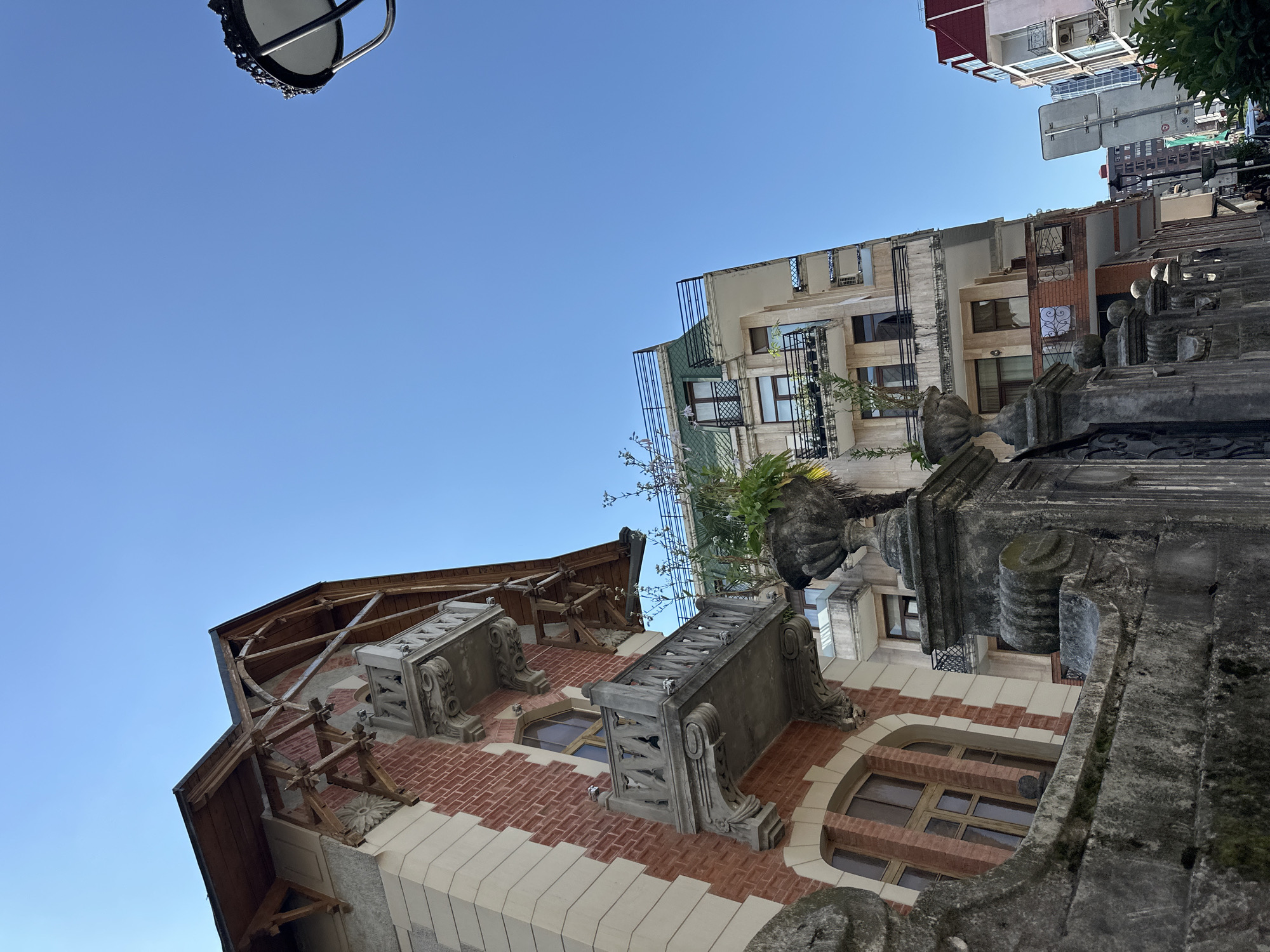 The image captures a low-angle view of a street scene in Batumi, Georgia, under a clear blue sky. In the foreground, a weathered, ornate stone fence and pillars are prominent. Behind the fence, a red brick building with decorative white trim and multiple balconies with intricate stone balustrades is visible. Adjacent to this building is a taller, modern apartment complex with a green roof and numerous balconies, some adorned with plants. Further down the street, other buildings of varying heights and architectural styles are present, along with a street lamp and a traffic sign. A few people can be seen walking in the distance, and a car is parked on the street. The scene appears to be a sunny day.